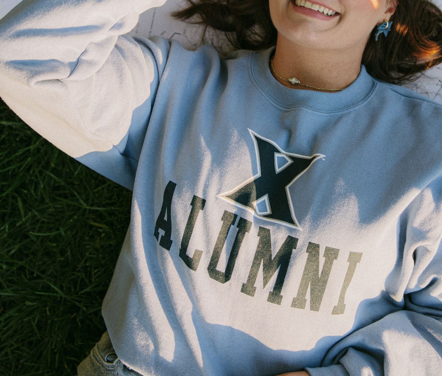 Smiling woman lying on grass wearing a light blue Xavier Alumni crewneck sweatshirt.