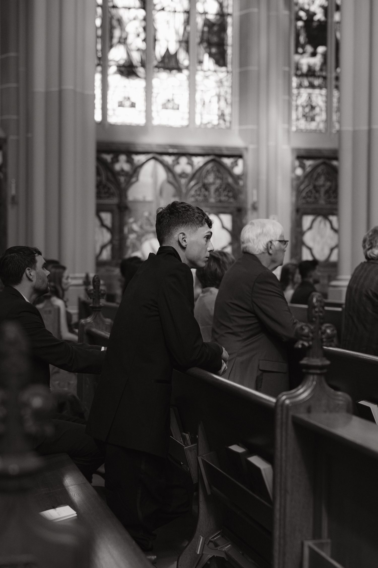 Black and white photo of wedding guests kneeling in prayer inside a Gothic cathedral with stunning stained glass windows.