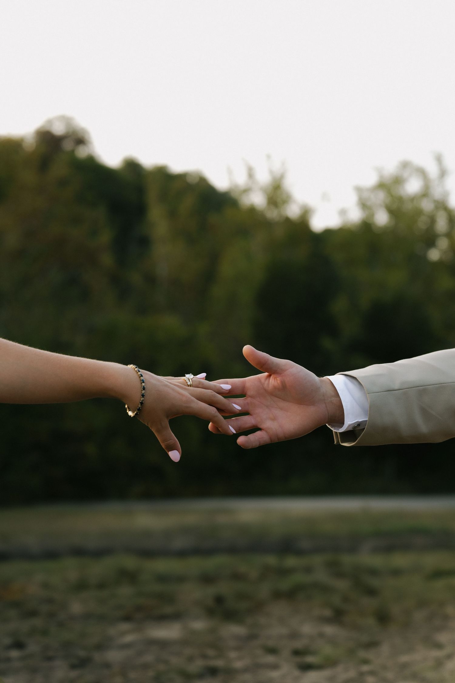 Bride and groom reaching toward each other hands with wedding ring and bracelet visible outdoors.