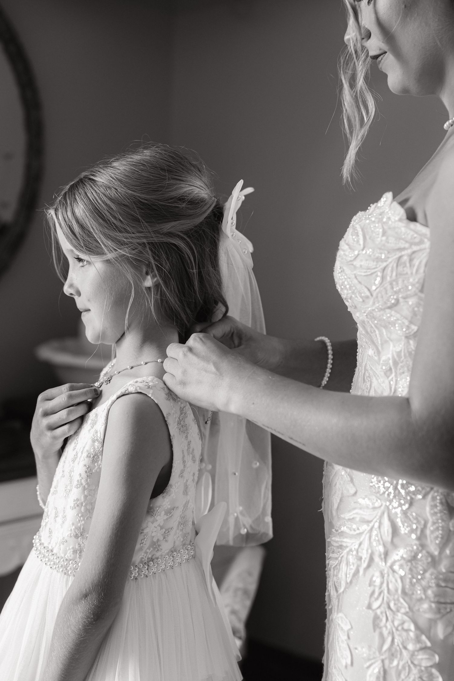 Black and white photo of bride in lace gown adjusting flower girls hair before wedding.