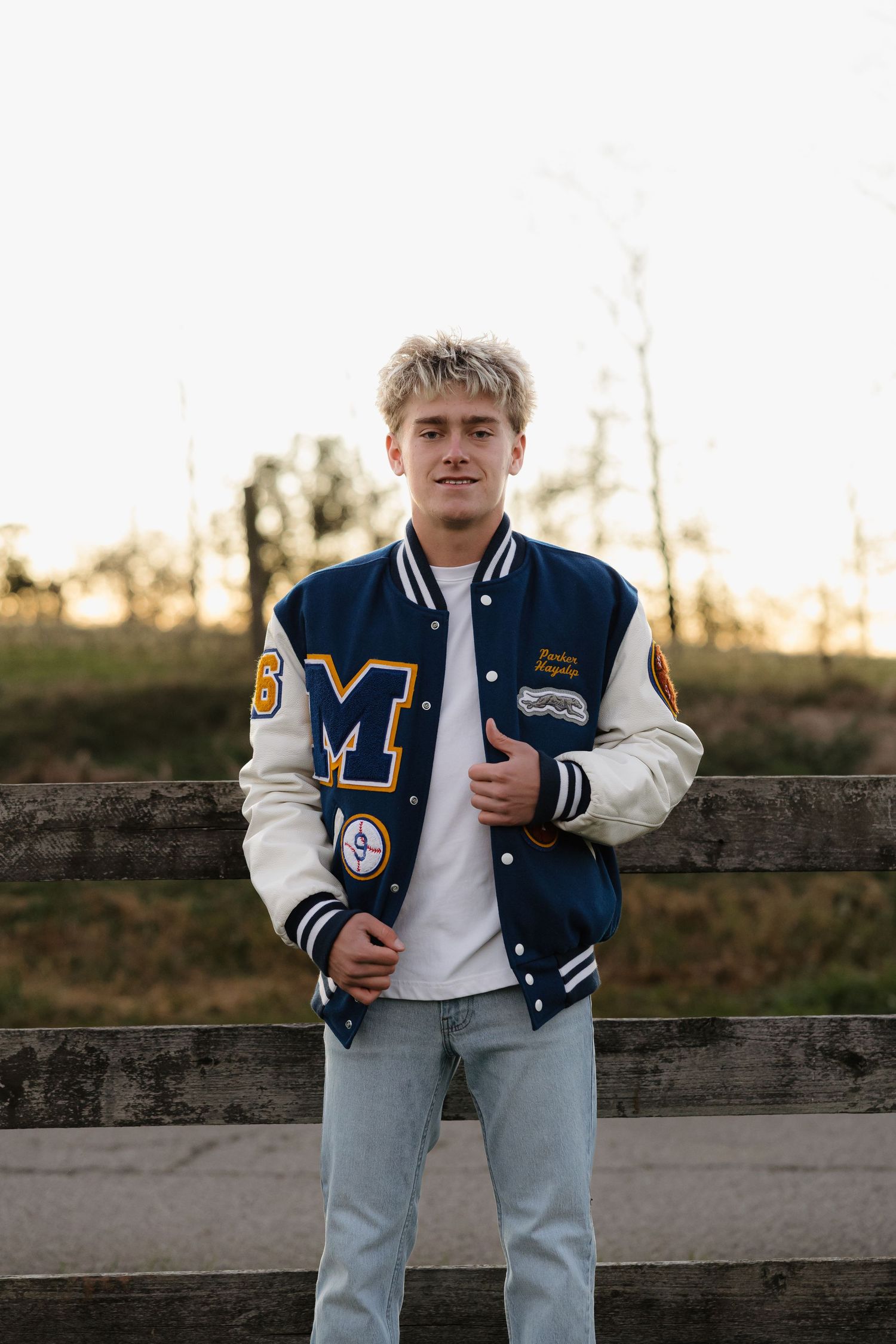 Teen boy in navy and white varsity letterman jacket posing by wooden fence for senior portrait.