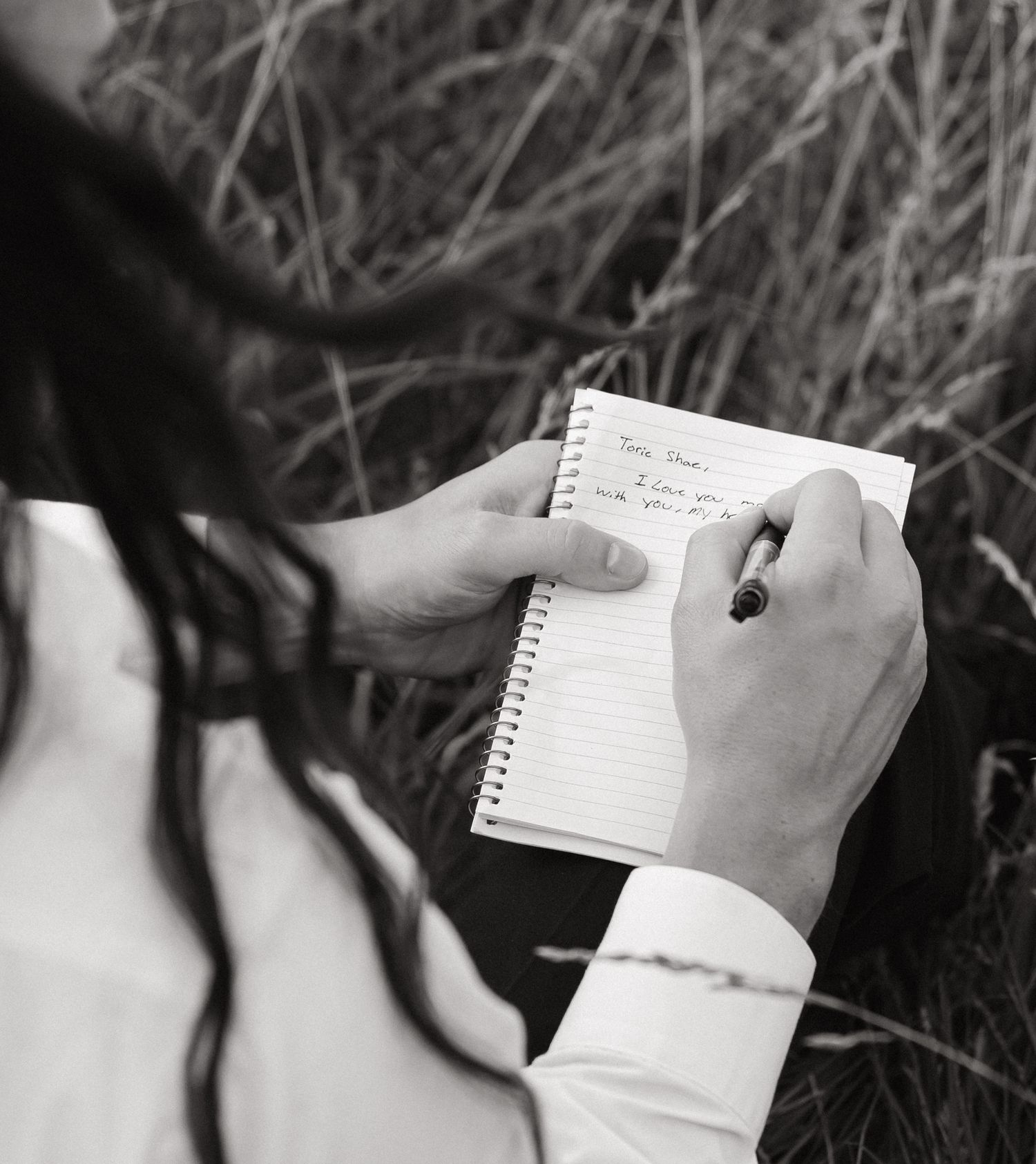 Black and white photo of a person writing a heartfelt note in a spiral notebook outdoors in a field.