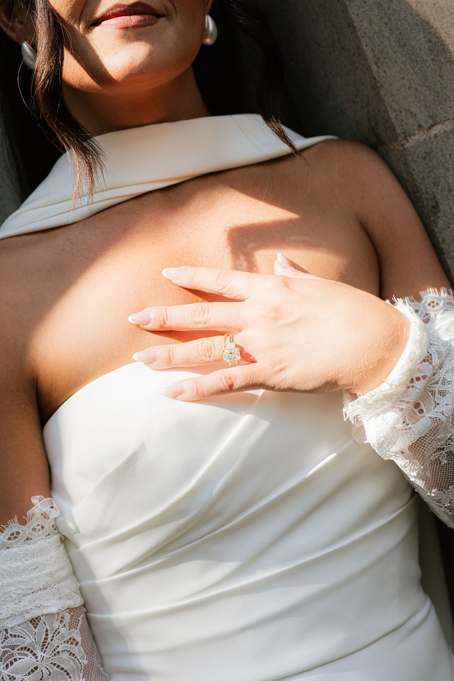 Bride in white strapless gown with lace sleeves showcasing elegant diamond engagement ring in sunlight.