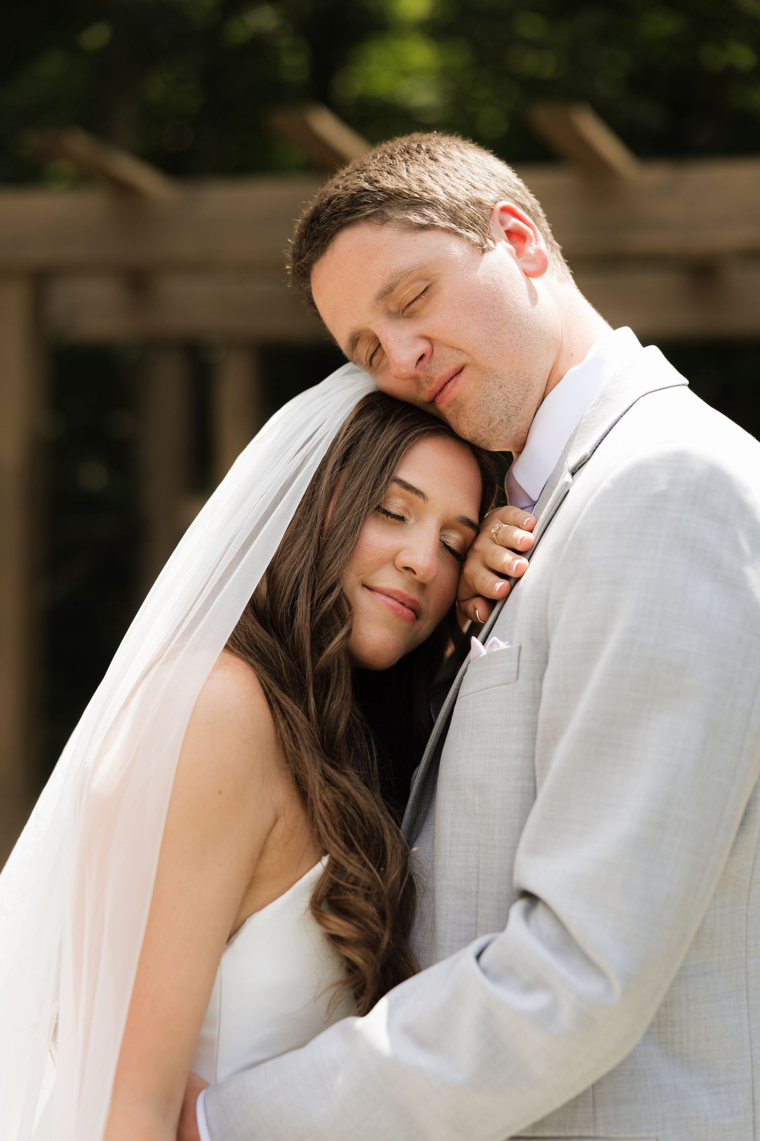 Bride and groom share a tender embrace, eyes closed, as she leans against his chest in wedding attire outdoors.
