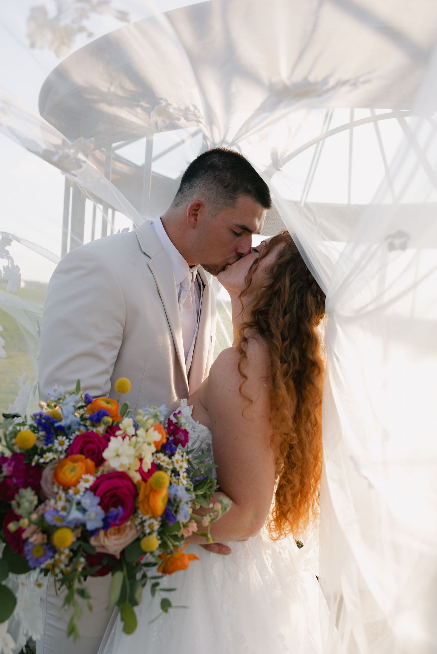 Bride and groom kissing with colorful bouquet and flowing veil outdoors.