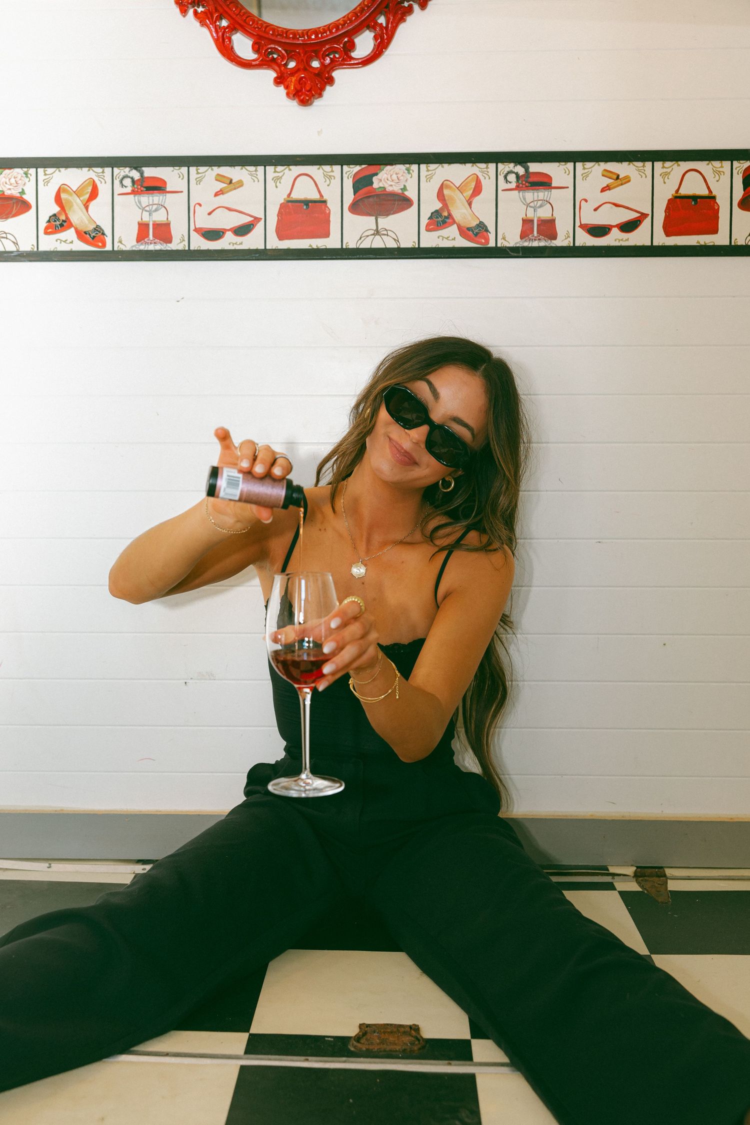 Stylish brunette woman in black outfit pouring red wine while sitting on checkered floor indoors.