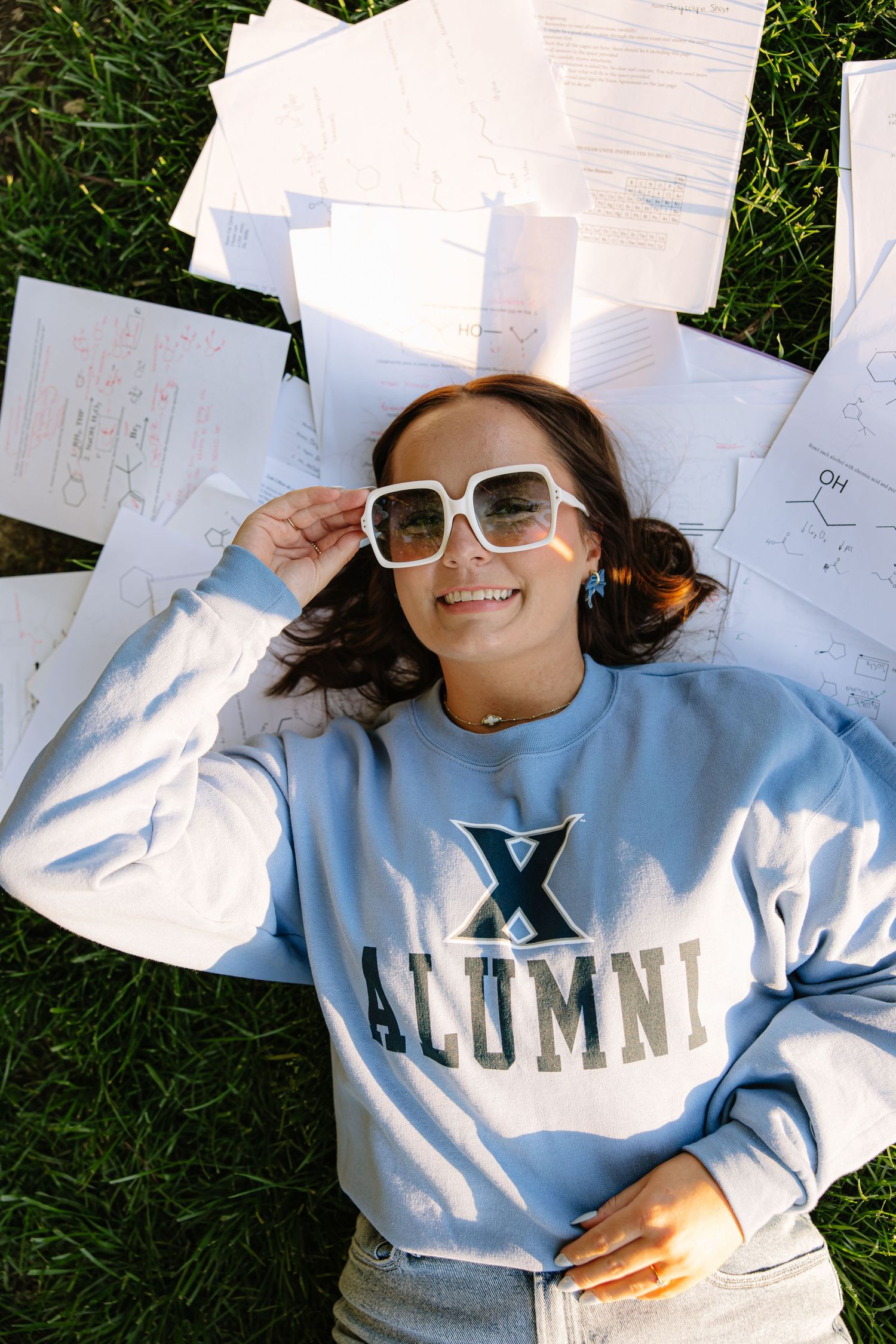 Smiling woman in Xavier Alumni sweatshirt lying on grass surrounded by study notes wearing white sunglasses.
