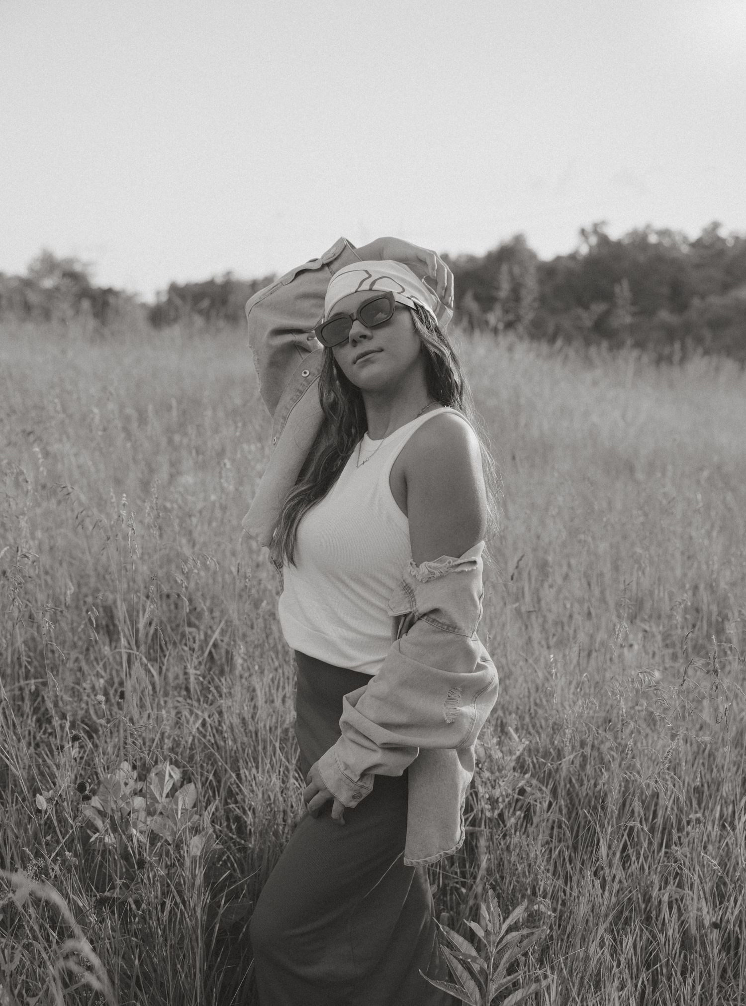 Black and white portrait of young woman in white tank top and sunglasses posing in tall grass field.