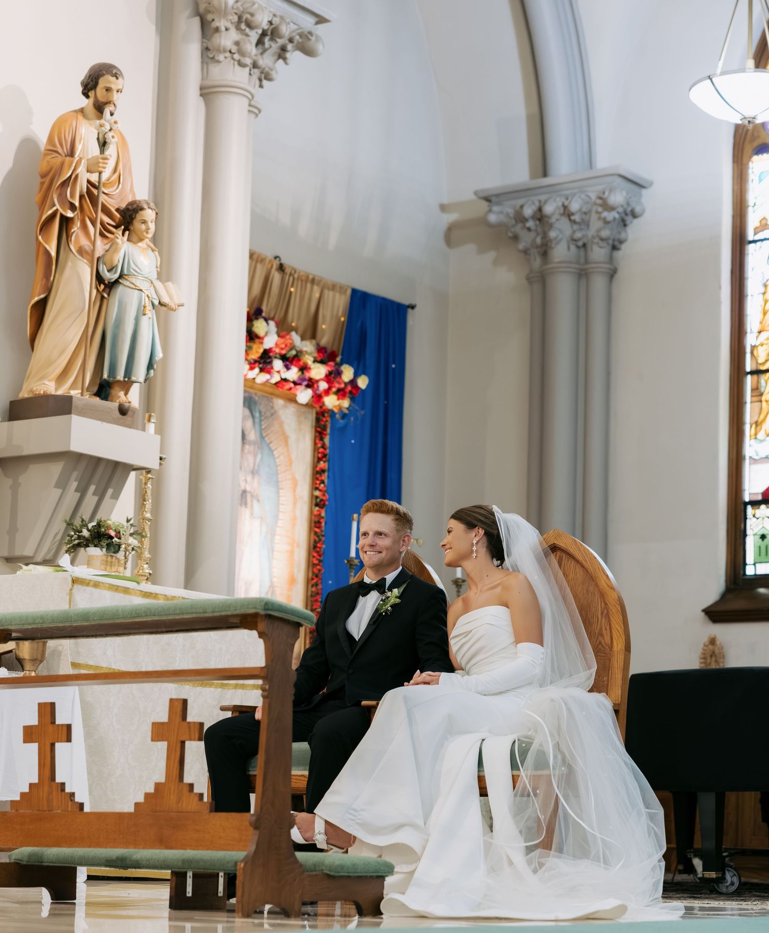 Bride and groom seated at ornate church altar with colorful floral arrangements during ceremony.