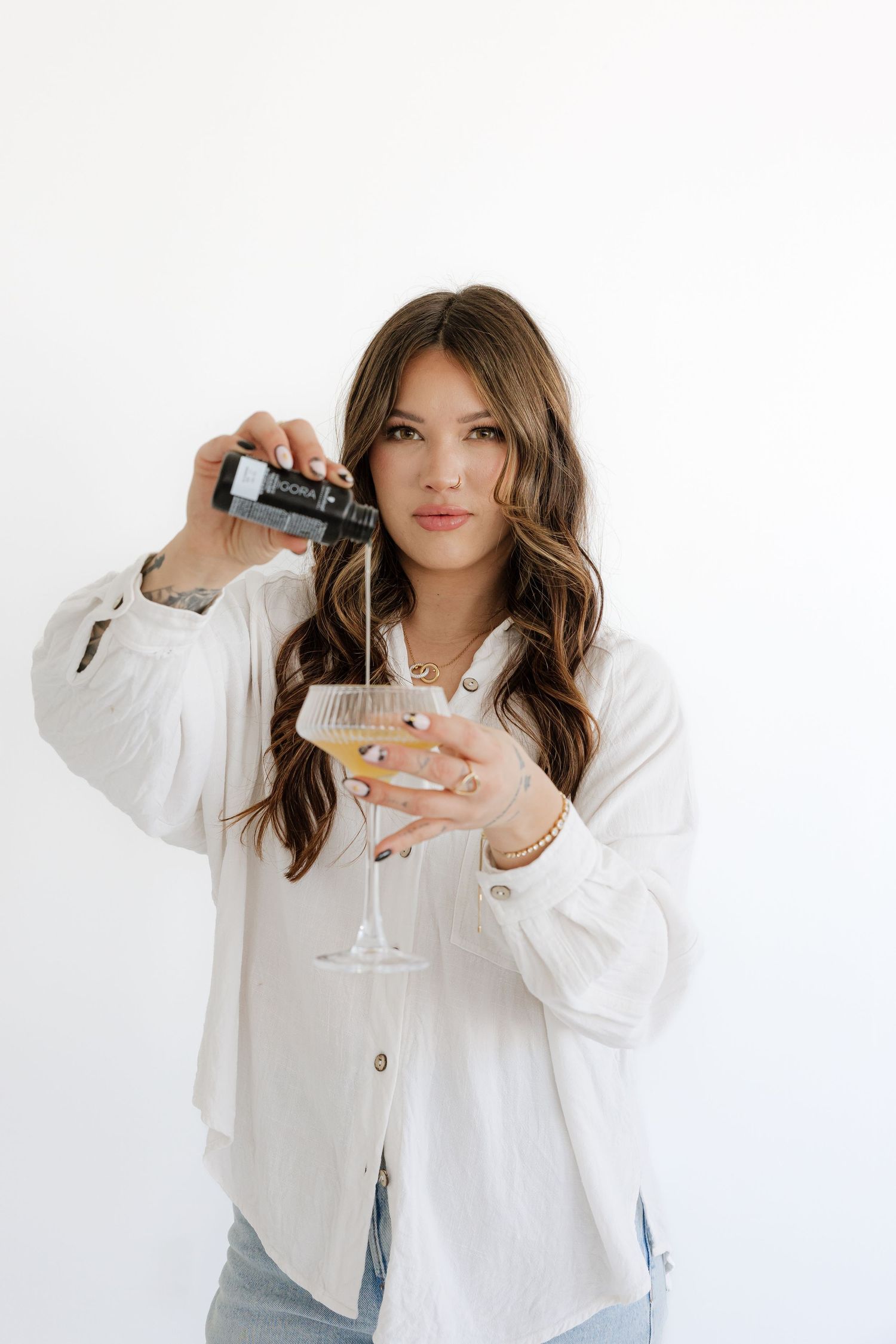 Stylish brunette woman in white shirt pouring a drink into a cocktail glass on white background.