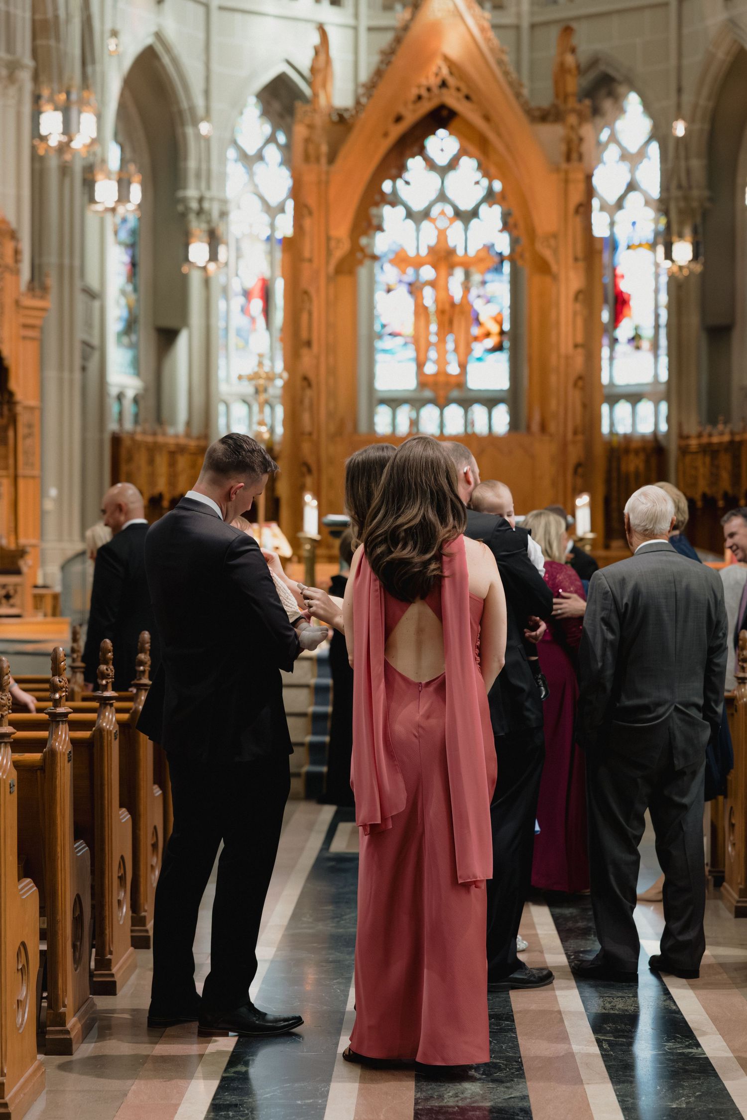 Wedding guests mingling in a grand Gothic cathedral aisle, woman in elegant backless rose-colored gown stands in foreground.