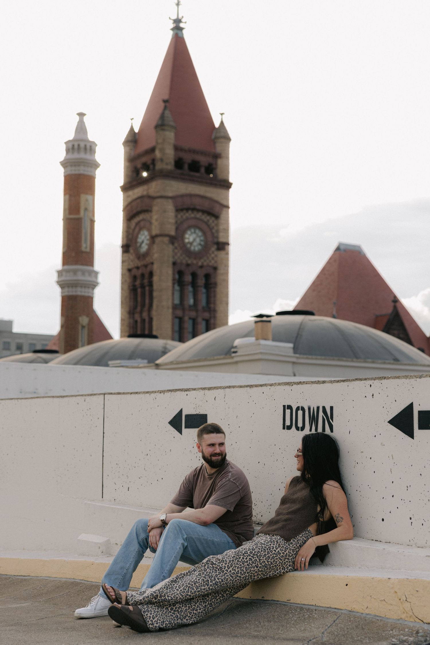 Couple sitting on rooftop ledge with historic clock tower building in background for engagement photos.