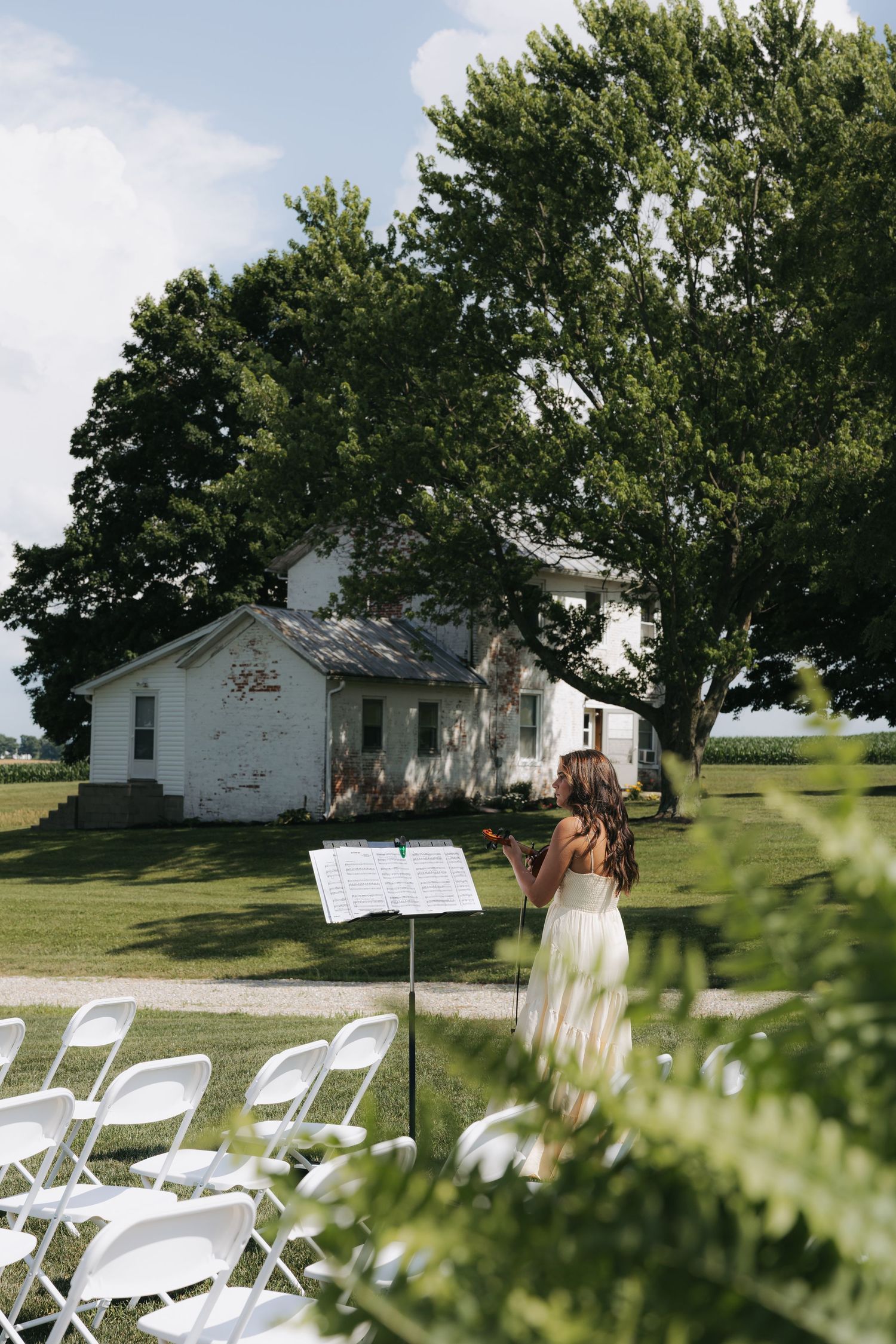 Violinist in white dress performing at an outdoor wedding ceremony near a rustic white farmhouse.