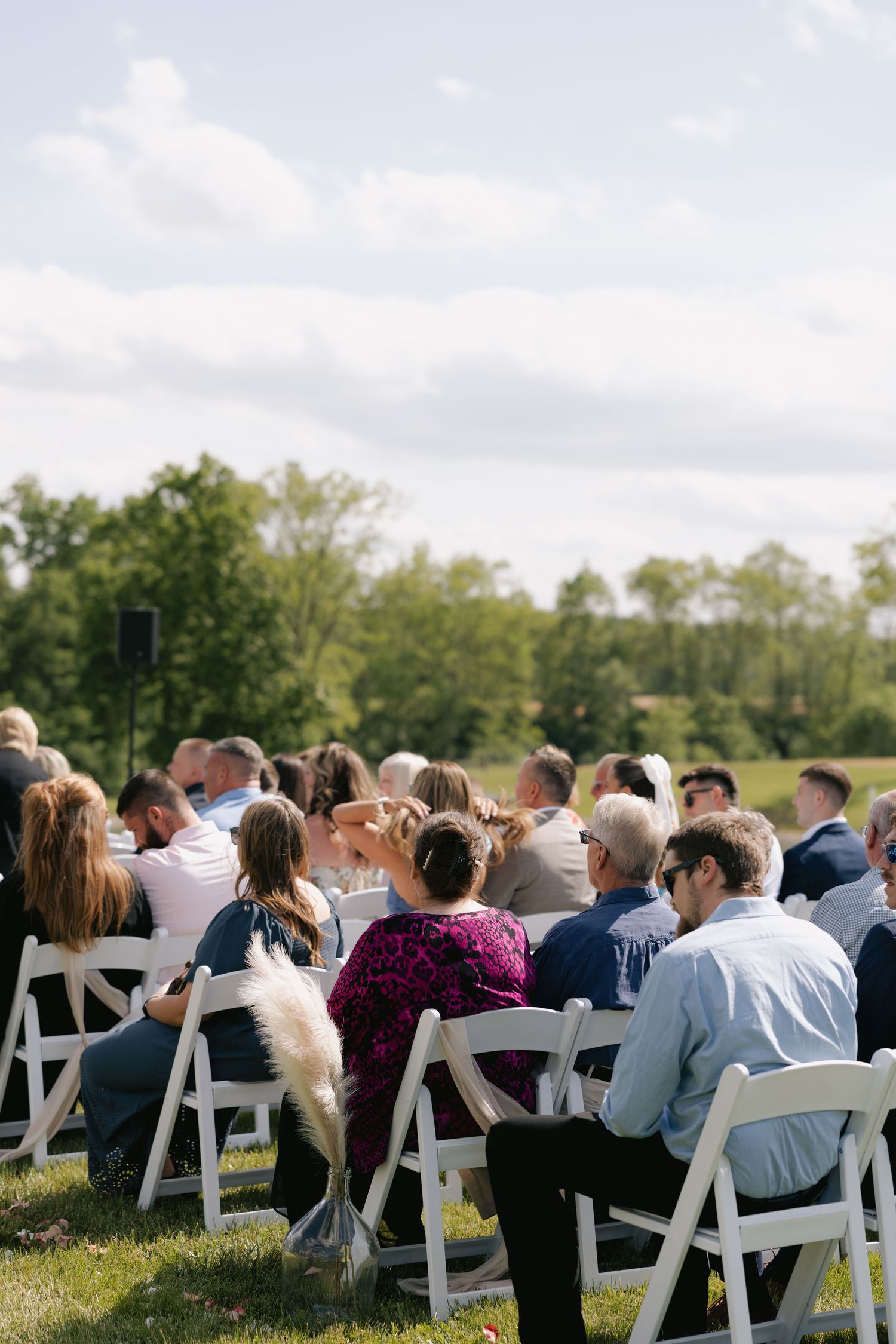 Wedding guests seated in white folding chairs at an outdoor ceremony on a sunny day.