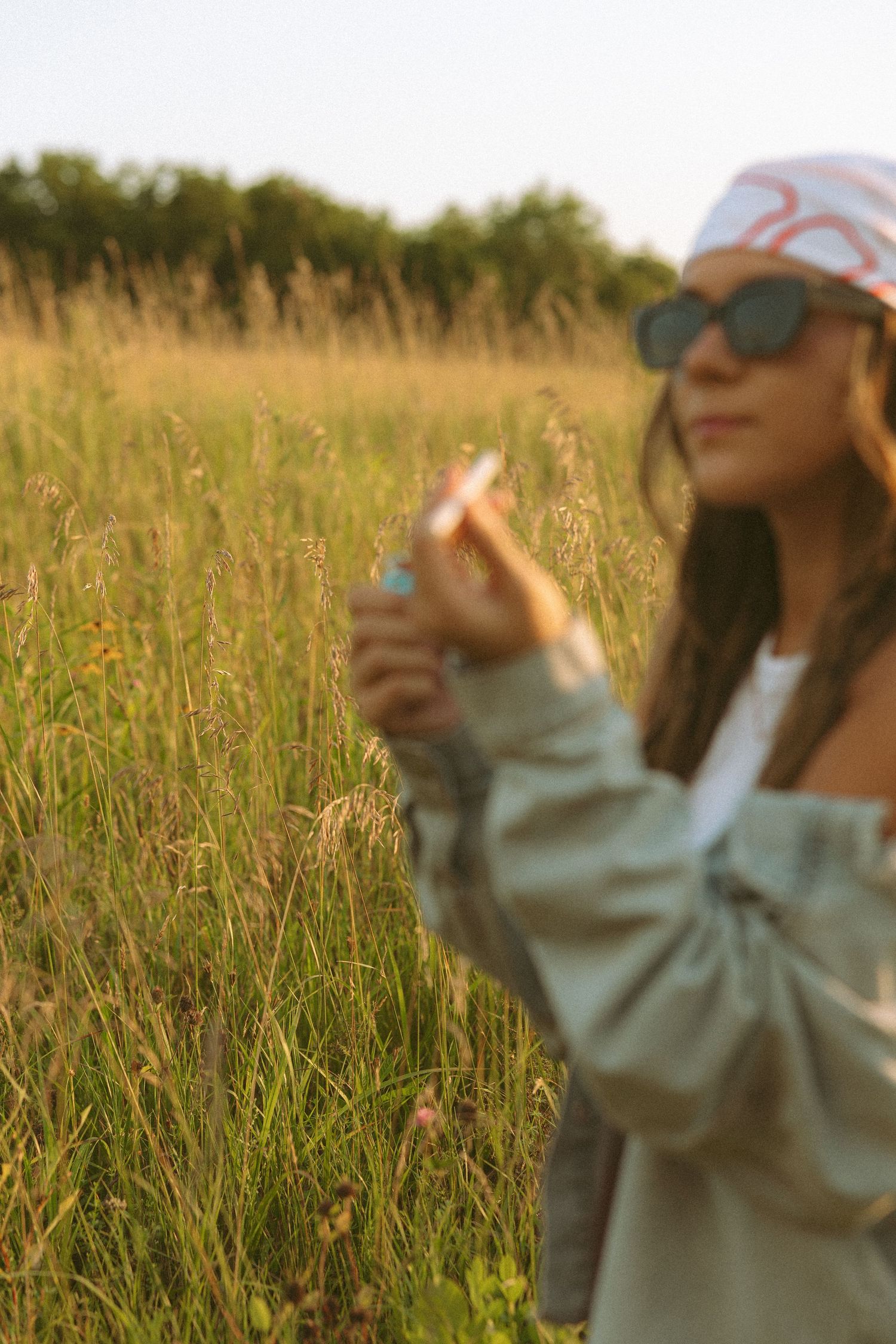 Blurred woman in sunglasses and bandana holding a cigarette in a golden sunlit tall grass field.