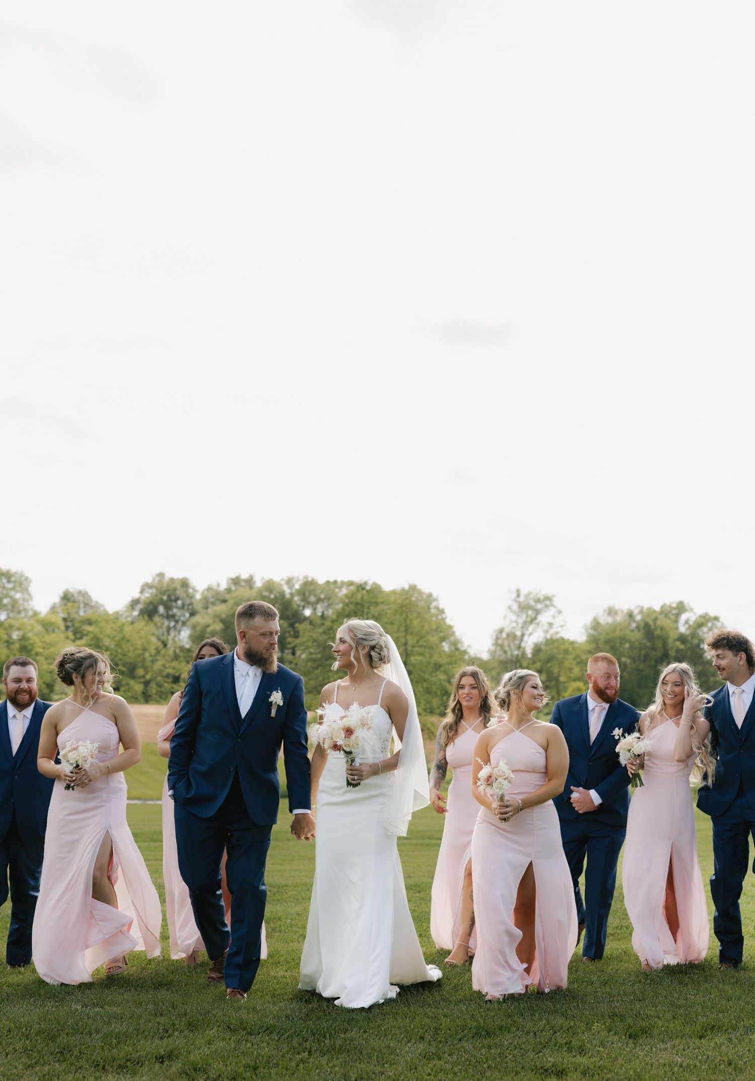 Bride and groom walking with wedding party in blush and navy attire on a green lawn.