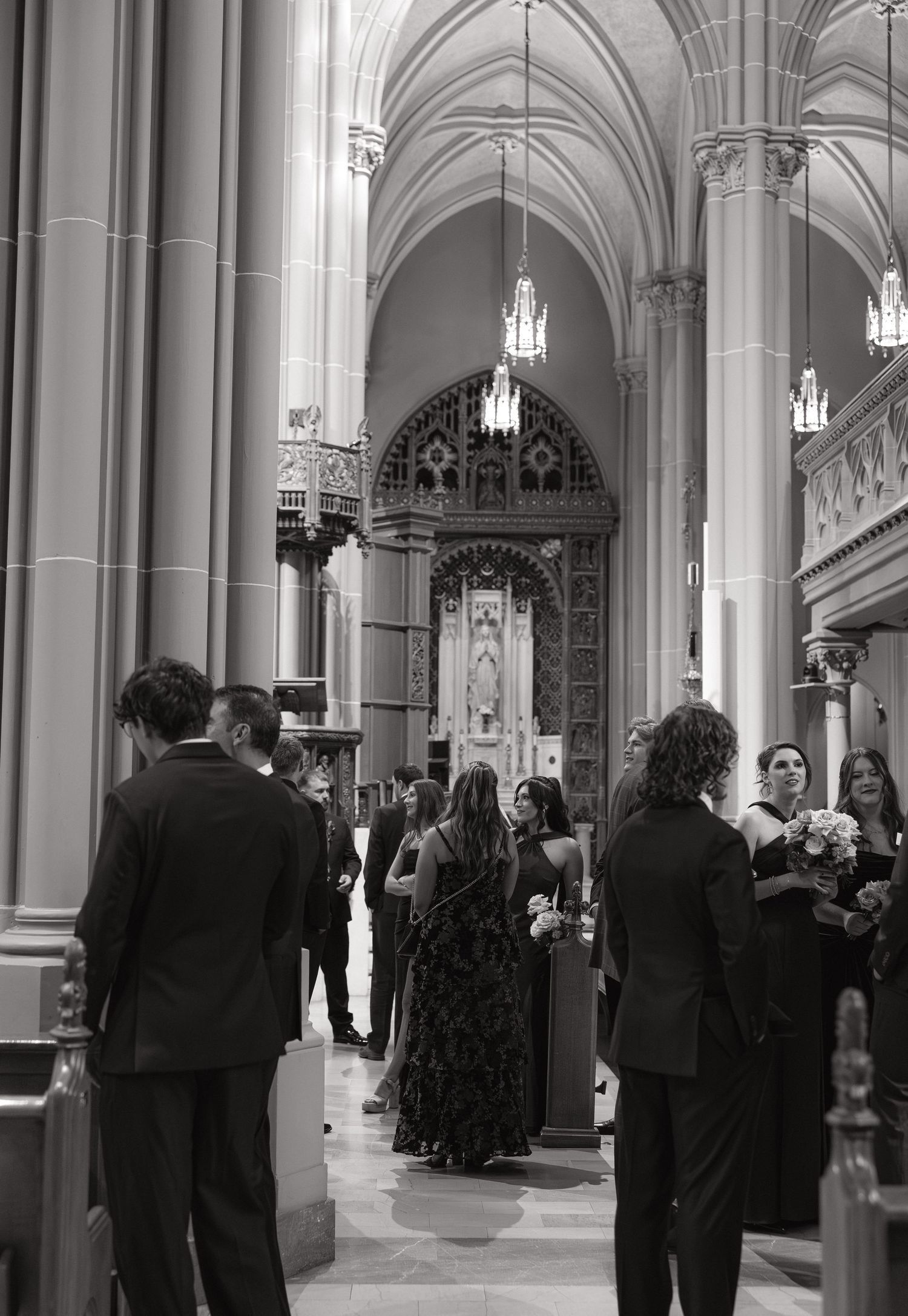 Black and white photo of wedding guests gathering in a majestic cathedral nave with towering Gothic arches and chandeliers.