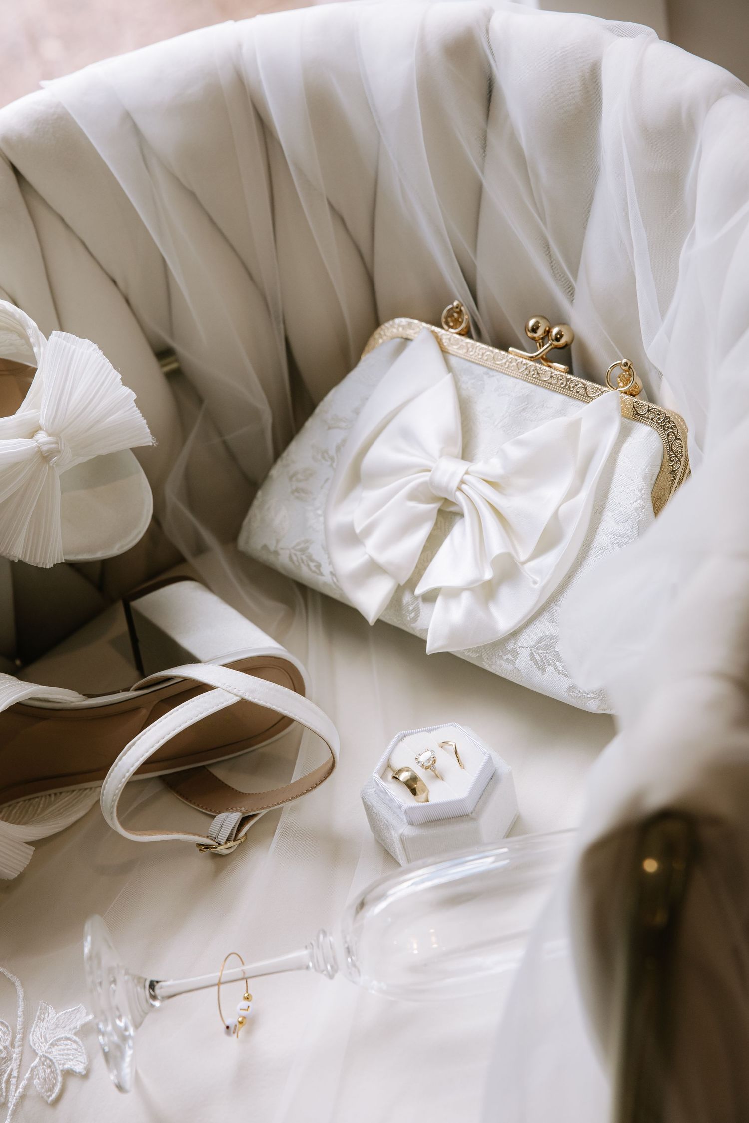 Elegant bridal accessories including white bow clutch purse, heeled sandals, gold rings in jewelry box, and sheer veil on chair.