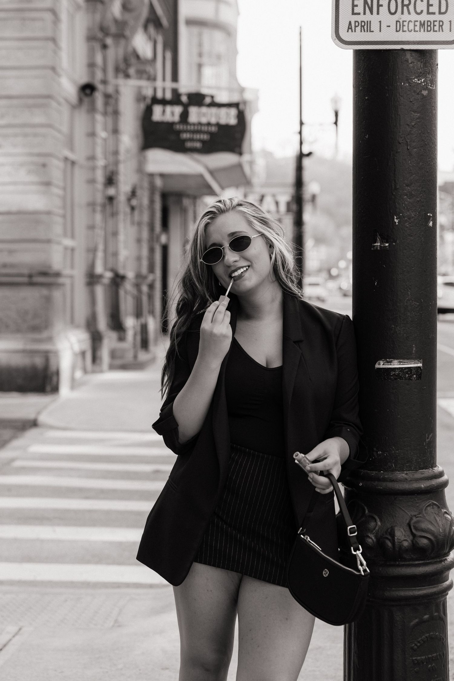 Black and white photo of smiling blonde woman in black blazer leaning on street pole in urban setting.