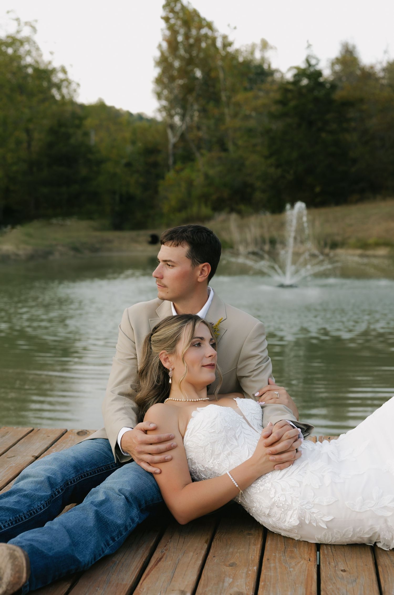 Bride and groom relaxing on a wooden dock by a pond with a fountain at sunset.