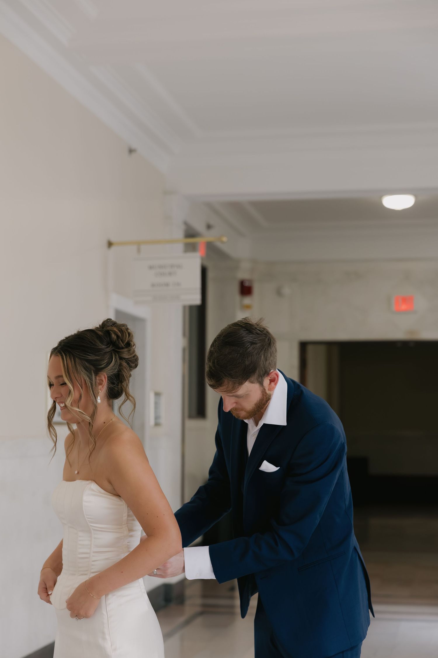 Groom in navy suit fastening bride's white strapless wedding dress in hallway before ceremony.