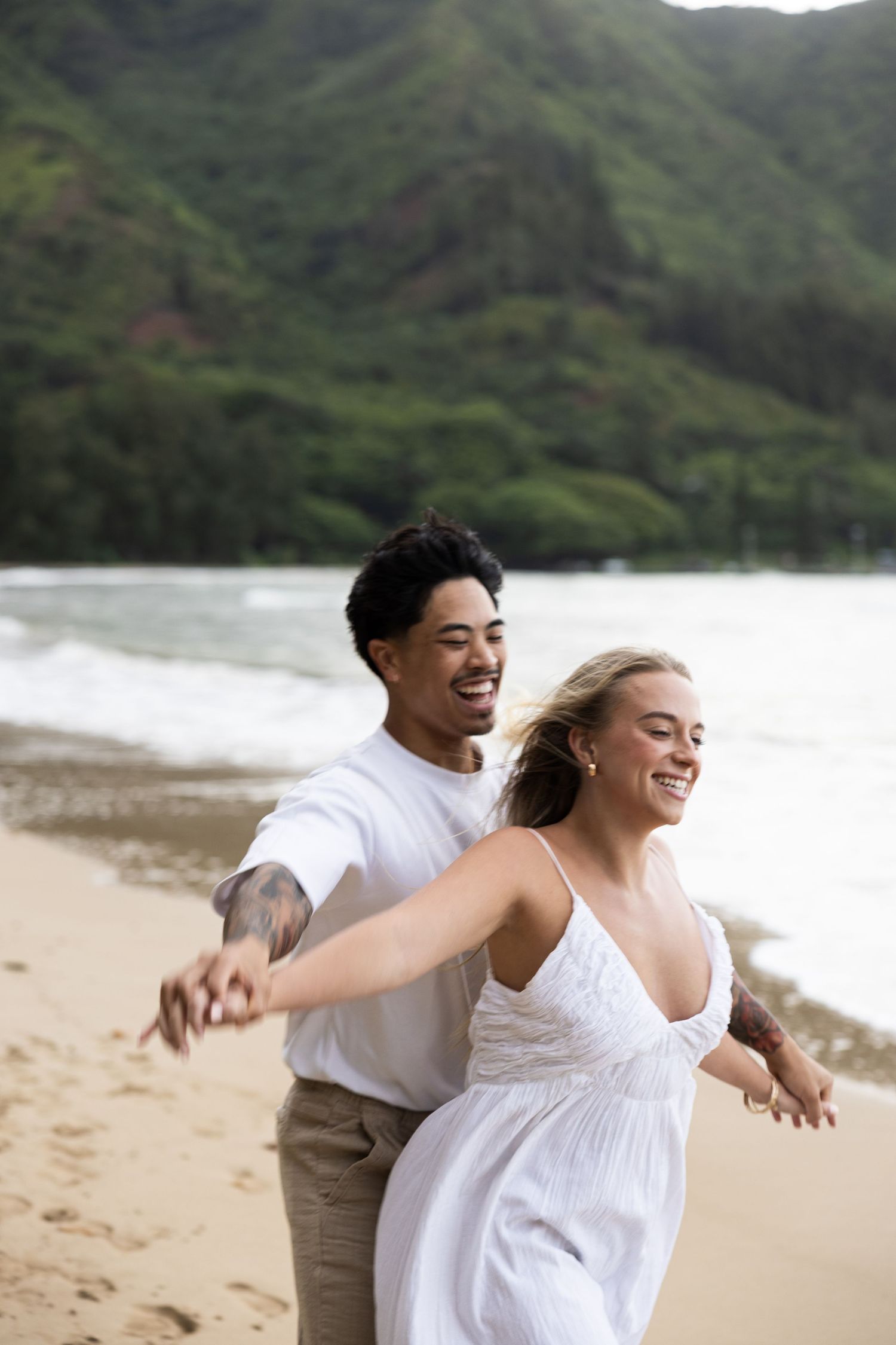 A joyful couple in white clothing shares a carefree moment together on a scenic beach with mountains in the background.