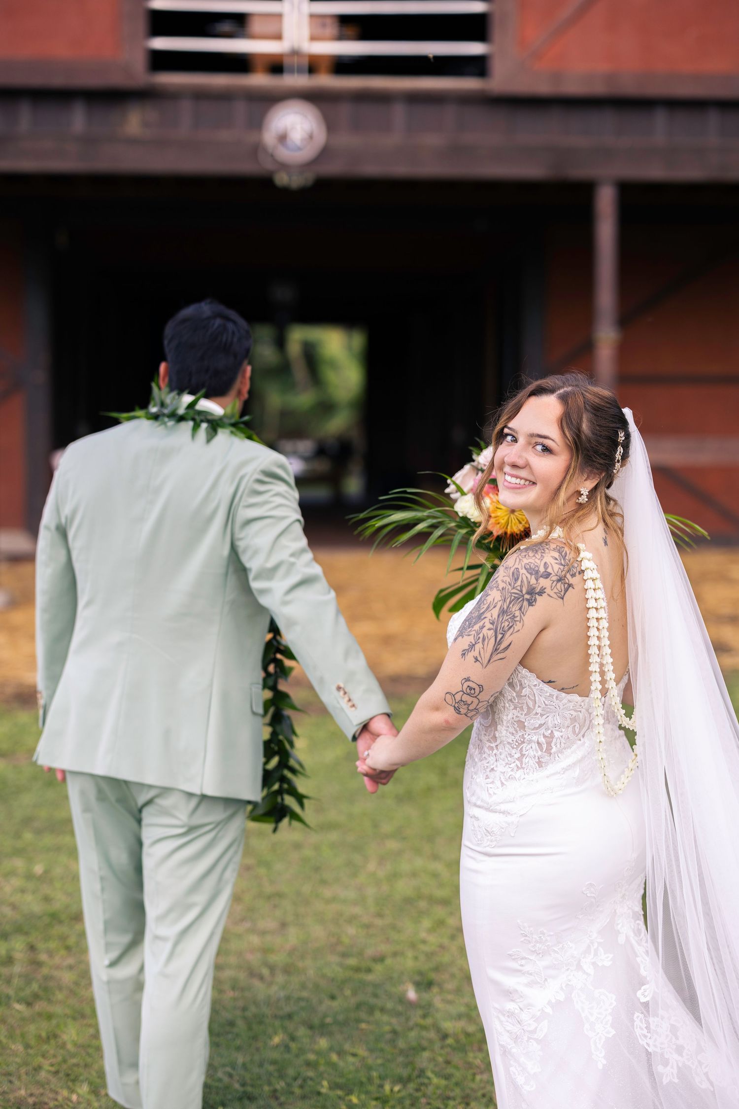 A bride in a lace wedding dress follows a groom in a mint green suit across a grassy outdoor venue.