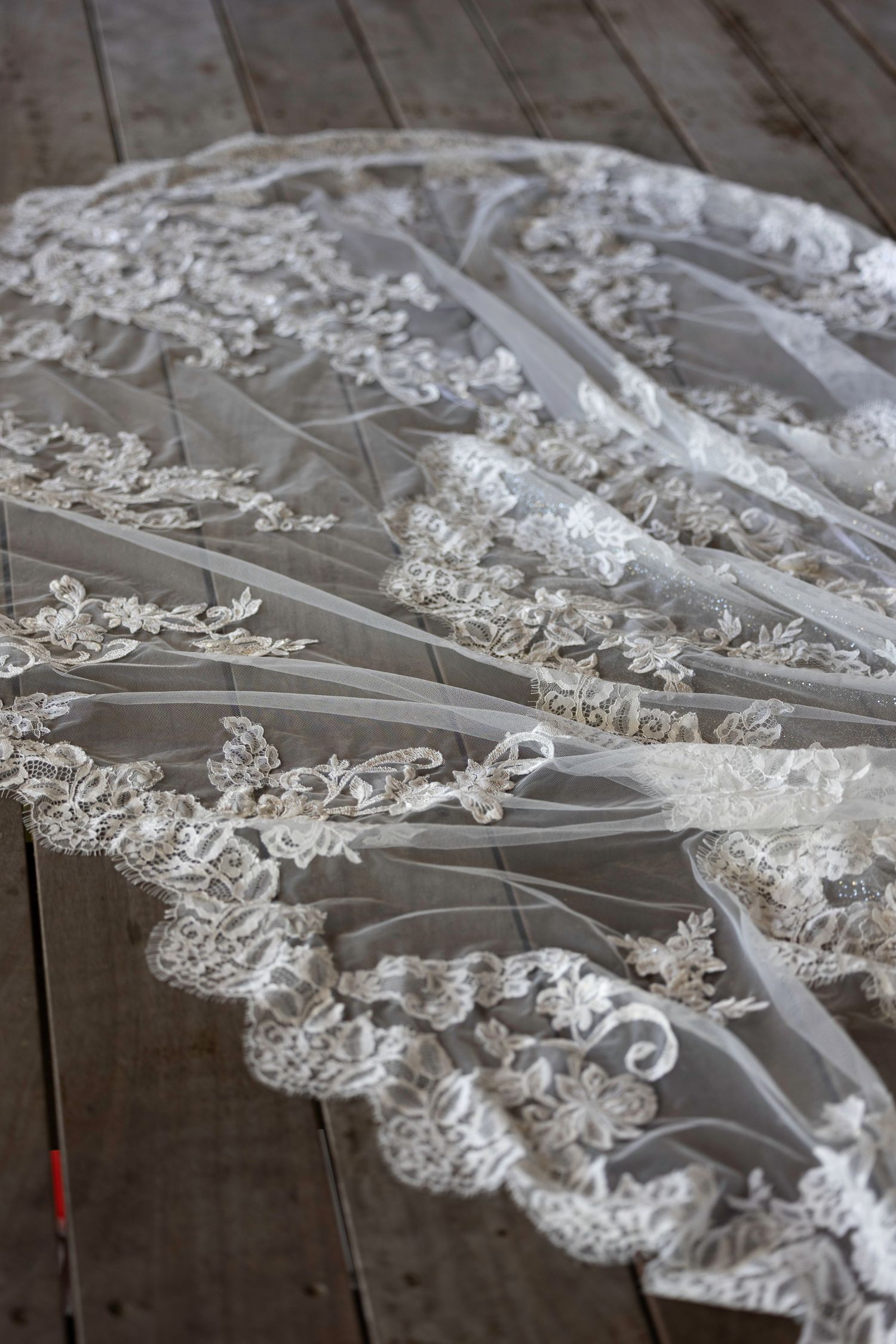 Delicate white lace bridal veil with intricate floral embroidery laid out on rustic wooden floorboards.