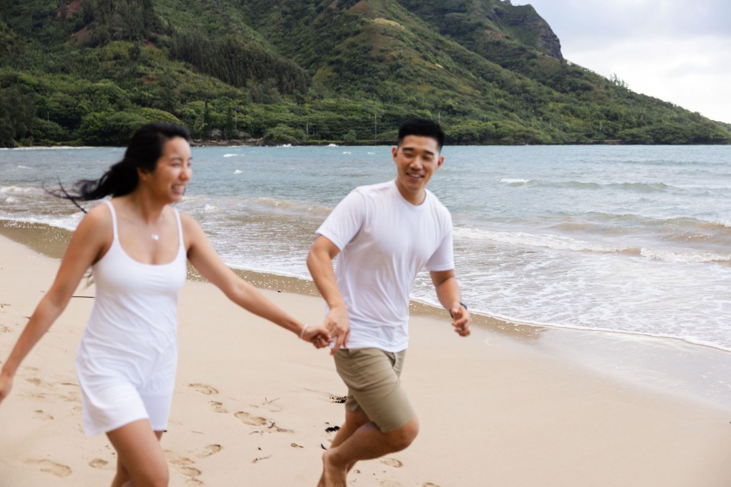 Two people playfully run together on a sandy beach with dramatic mountain cliffs in the distance.