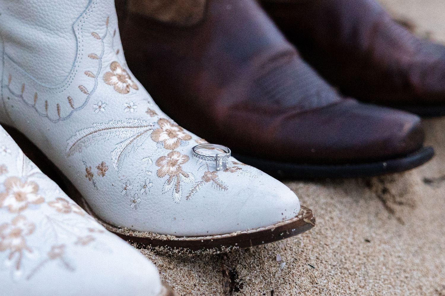 Close-up view of white leather cowboy boots with floral embroidery details sitting on sandy ground.