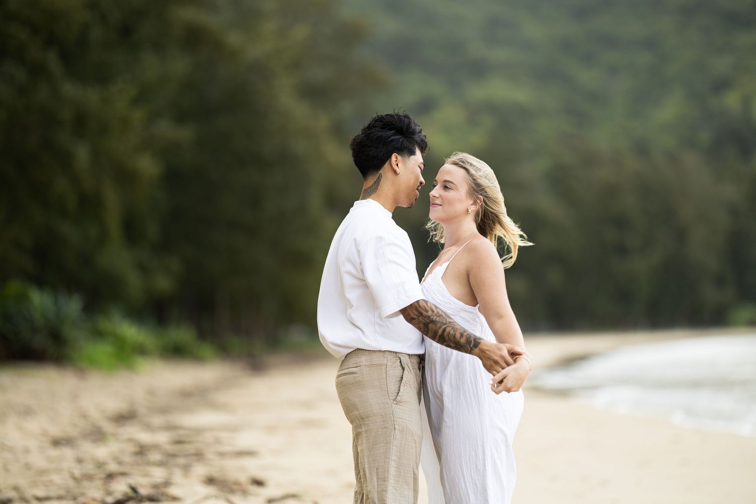A couple poses romantically on a beach with lush mountains in the background.