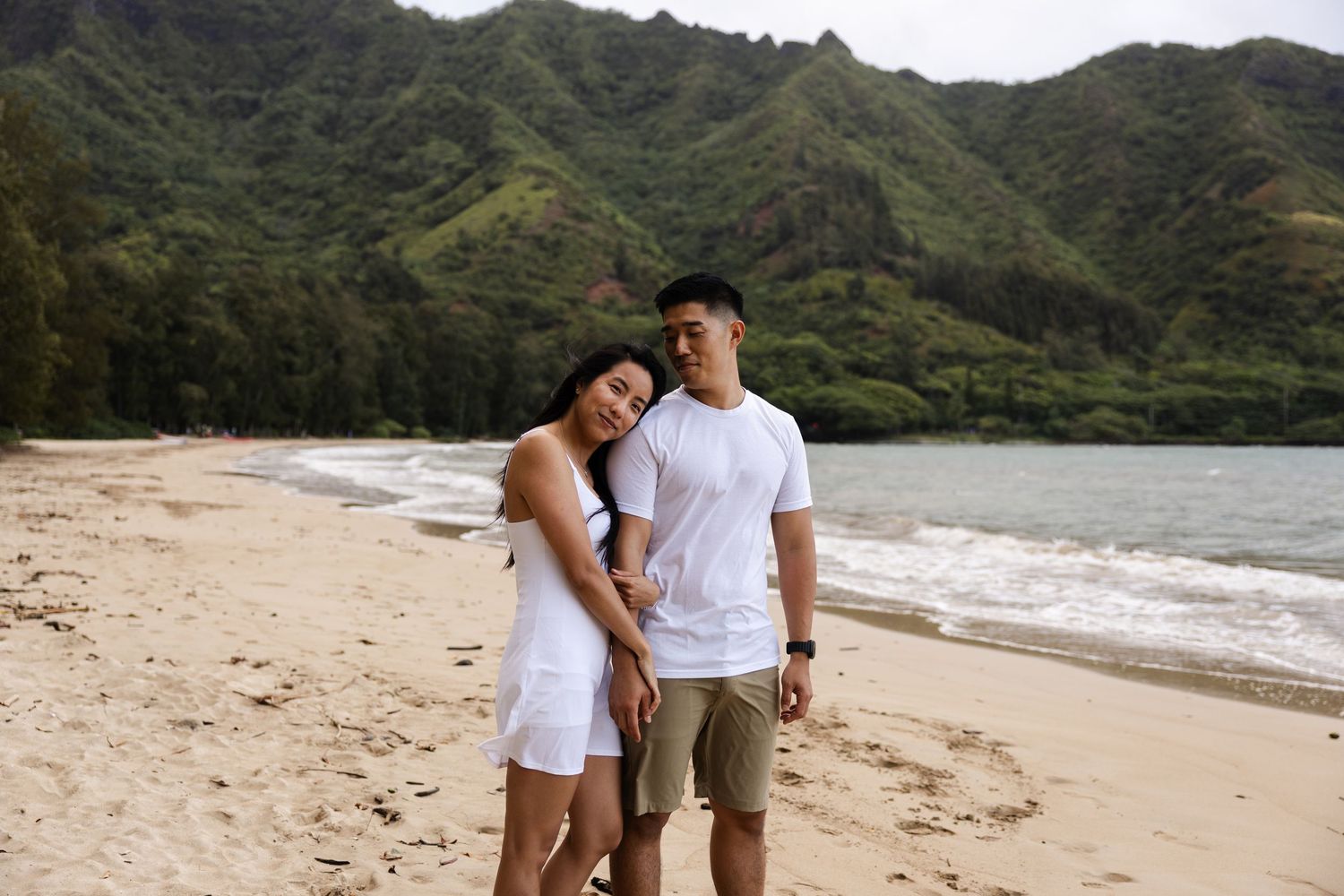 A couple shares a tender moment while walking on a tropical beach with verdant mountains overlooking the shore.