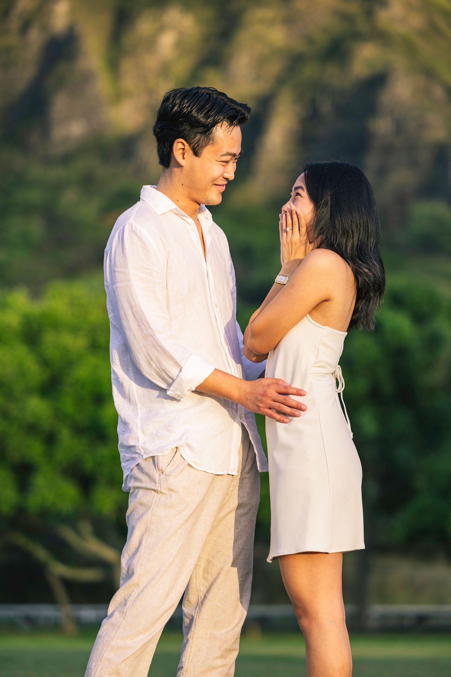 A couple in white outfits shares a tender moment together in a lush green outdoor setting during golden hour.