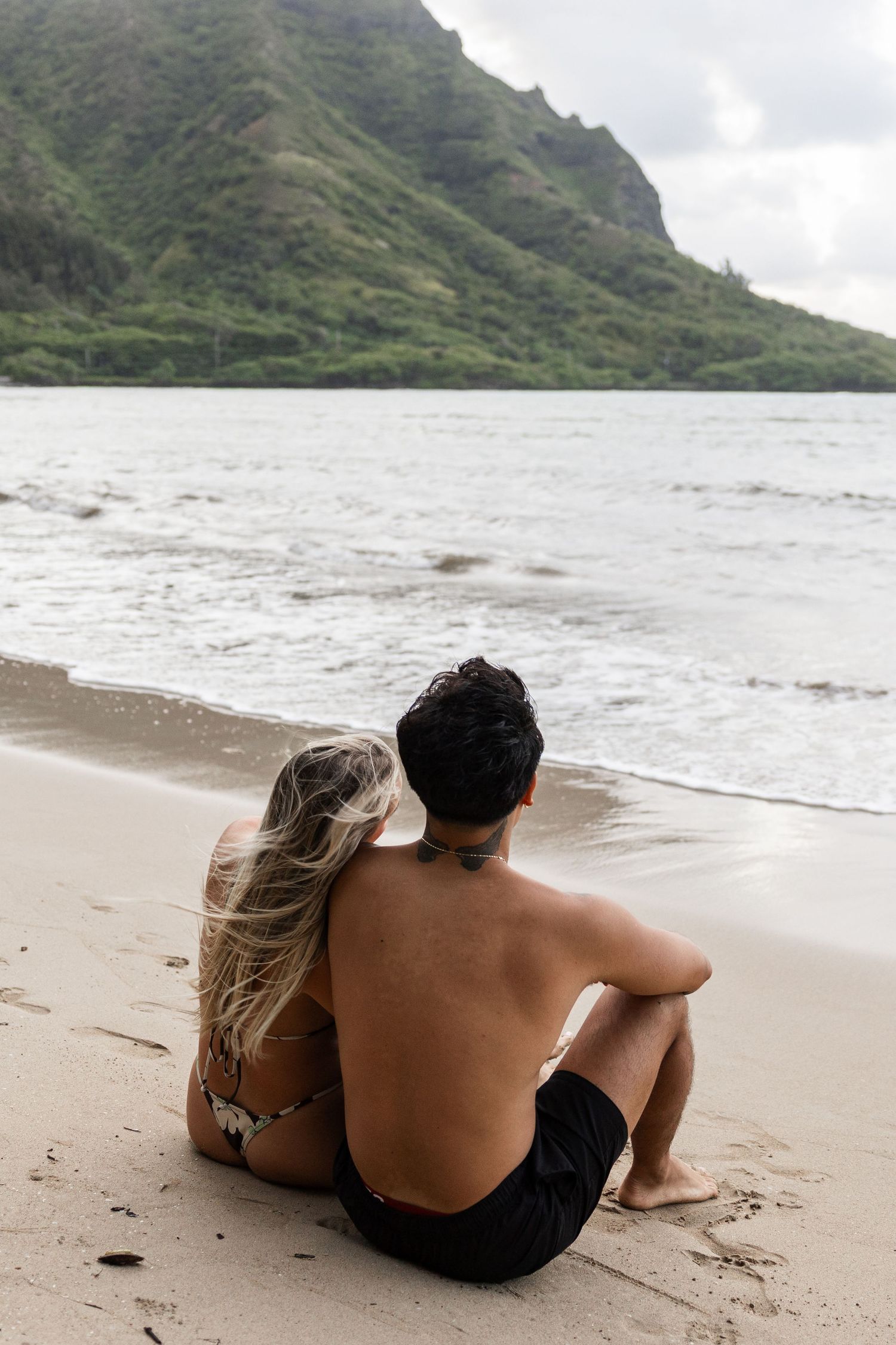 A couple sits together on a sandy beach overlooking dramatic mountain cliffs and ocean waves in Hawaii.