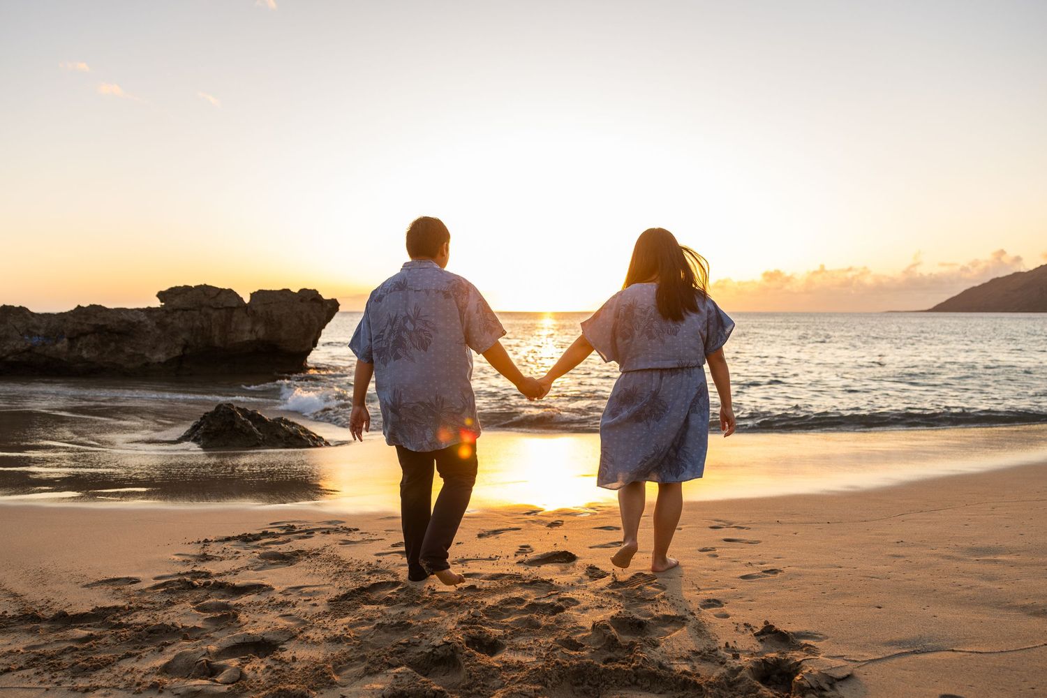 Two people walking hand in hand along a beach at sunset with waves crashing on the shore.