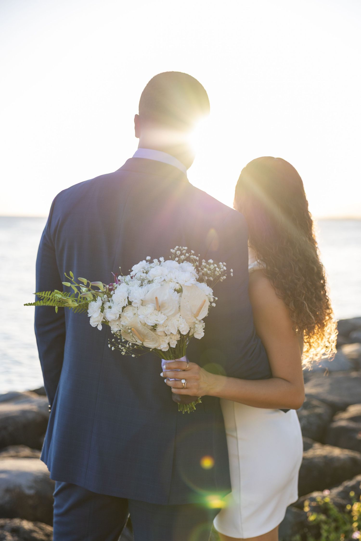 Backlit silhouette of newlyweds holding white bouquet against ocean sunset.