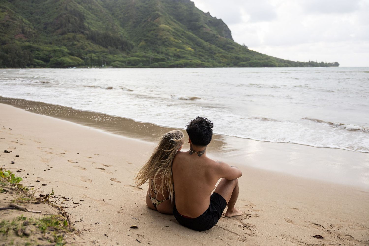 Two people sitting together on sandy beach watching waves with mountains in background.