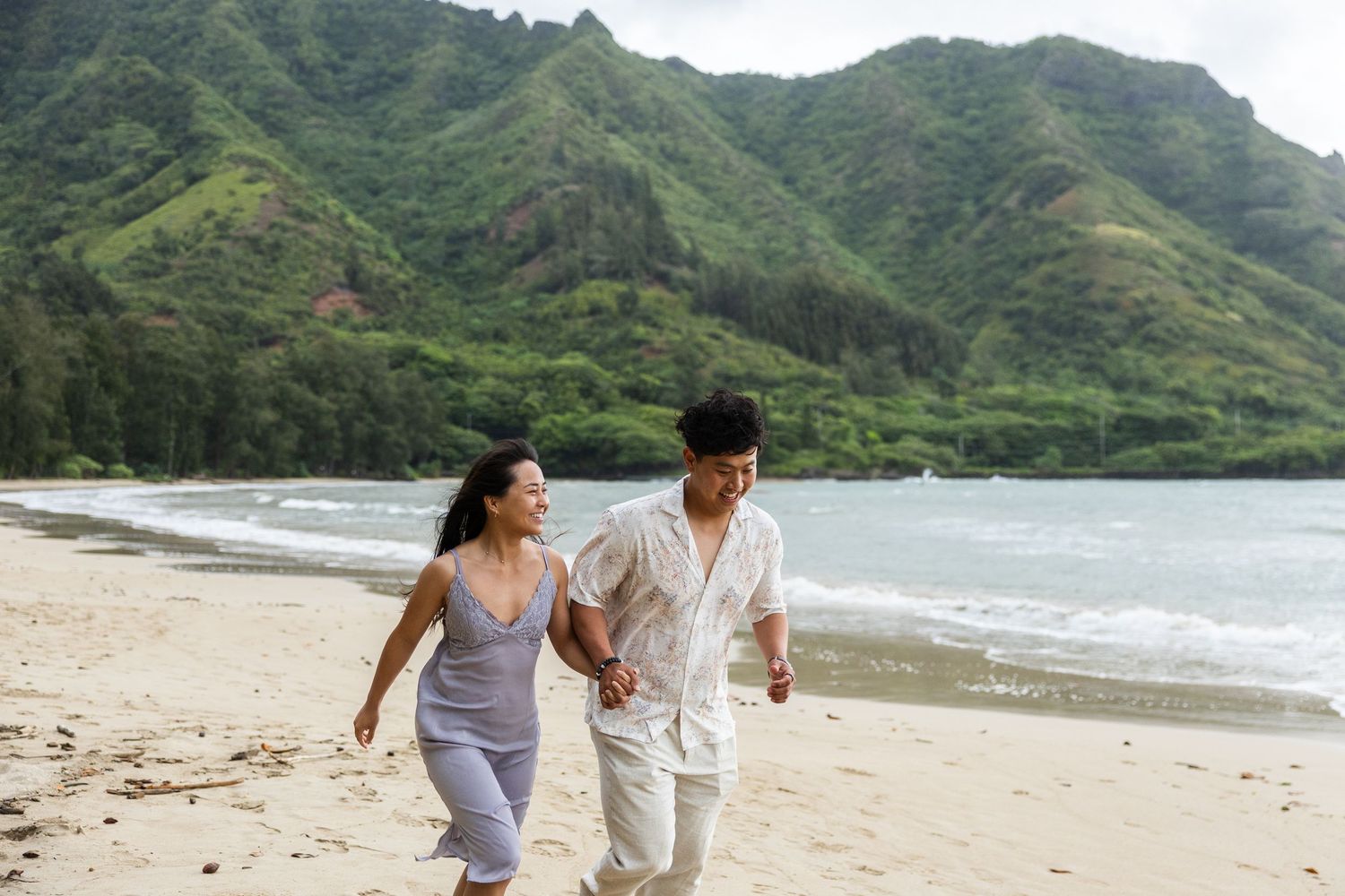 A couple enjoys a romantic walk along a scenic tropical beach with lush green mountains in the background.