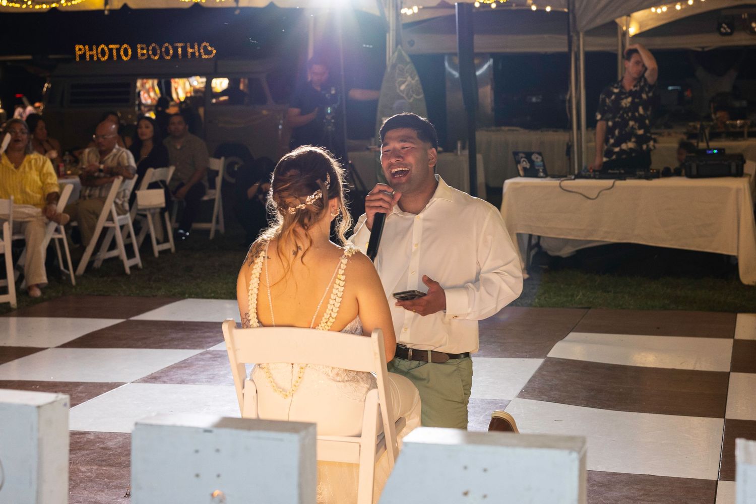 People gather around a photo booth setup at an outdoor nighttime event with string lights overhead.