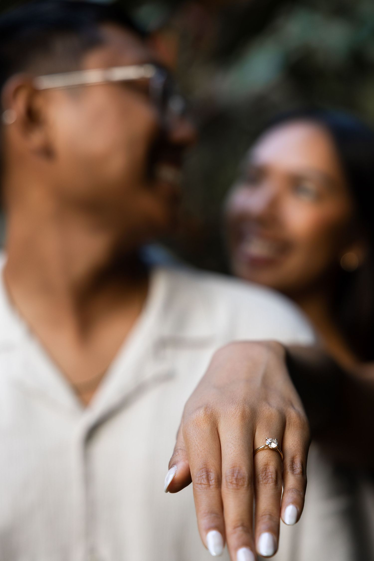 An engagement ring sparkles on a hand while a couple shares a happy moment.
