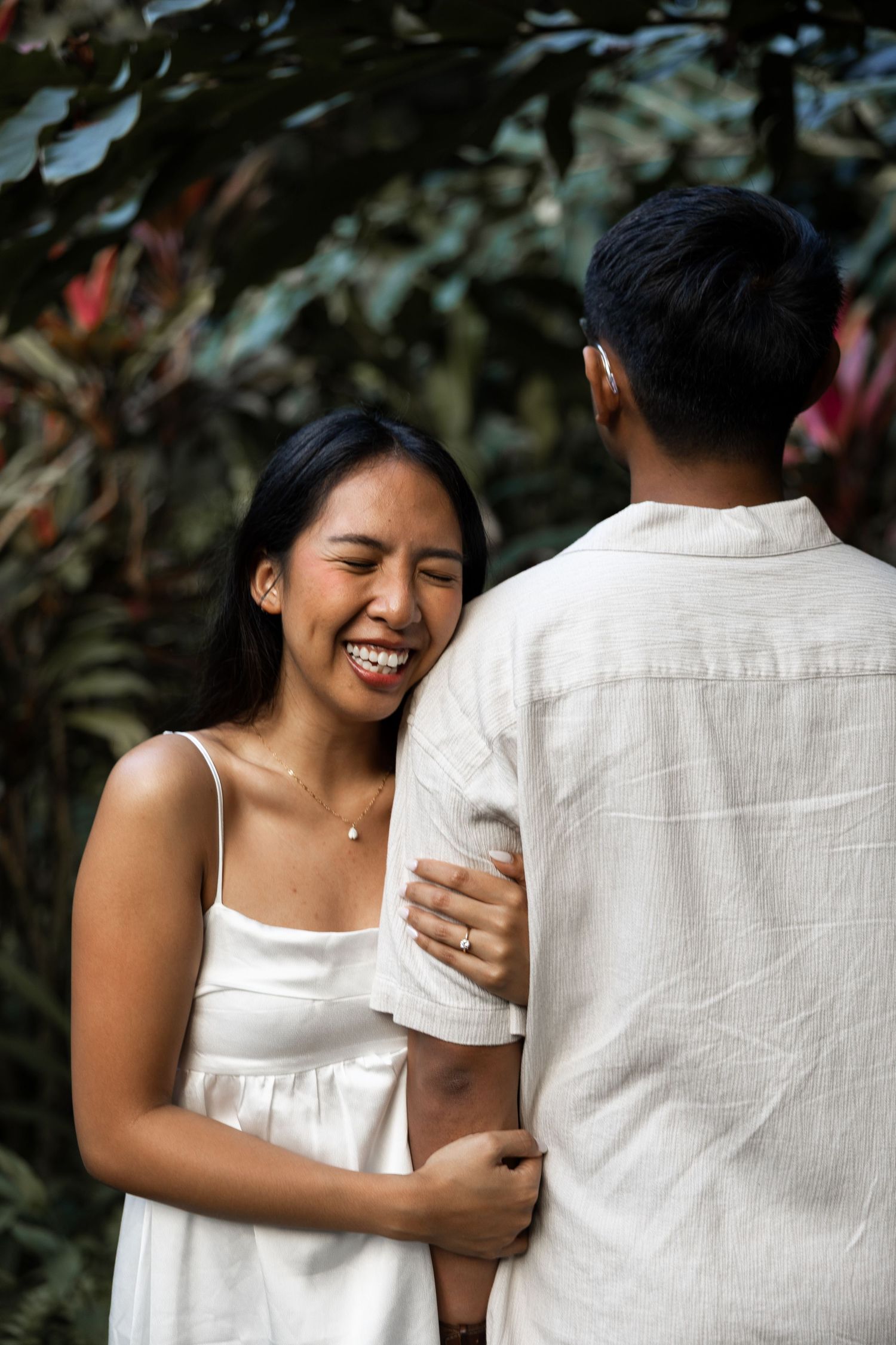 A joyful couple embraces in a garden setting wearing white casual outfits.