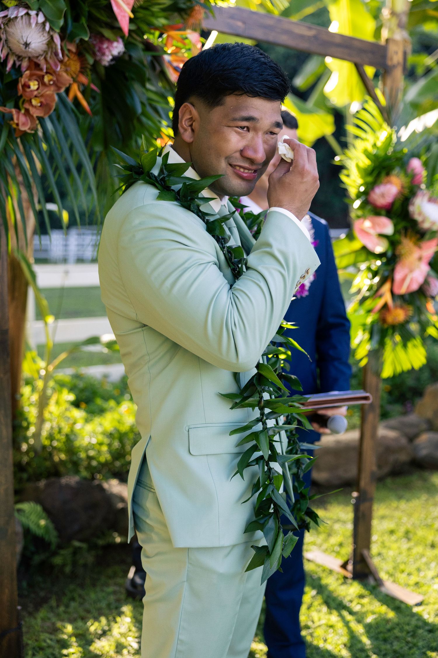Groom wearing a light beige suit and traditional Hawaiian lei stands outdoors during a wedding ceremony.