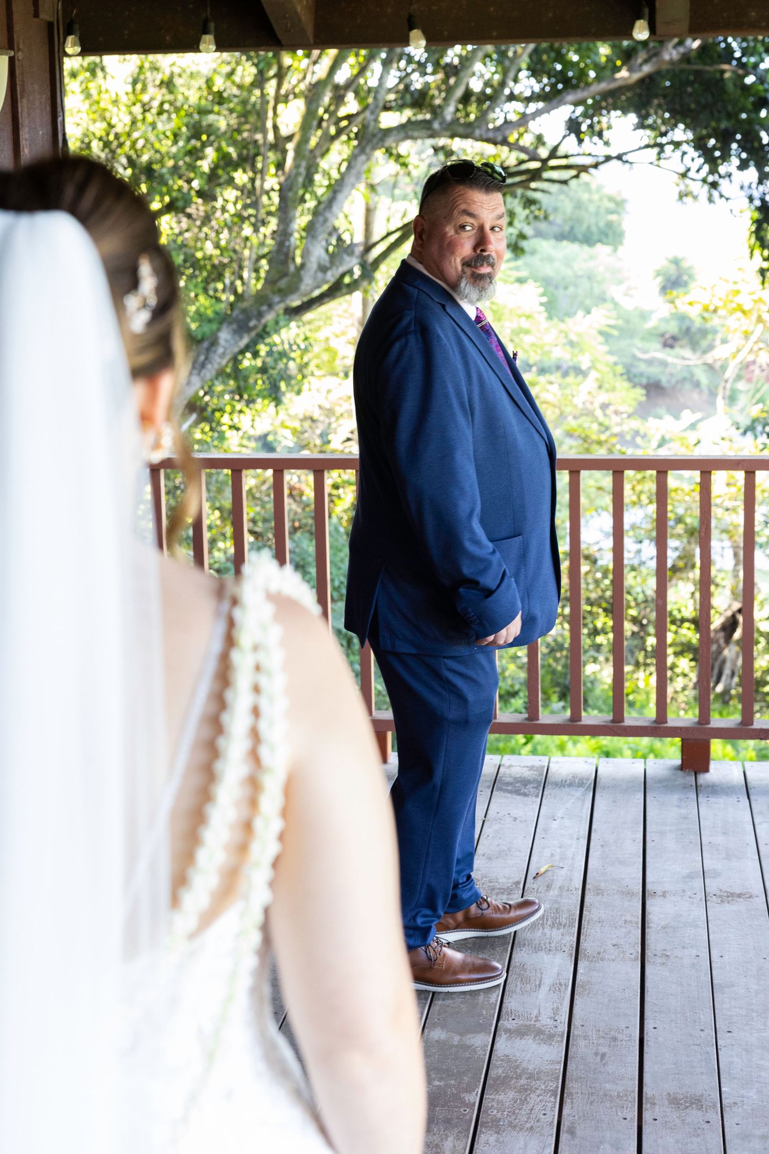 A person in a navy blue suit stands on a wooden deck surrounded by trees and natural lighting.