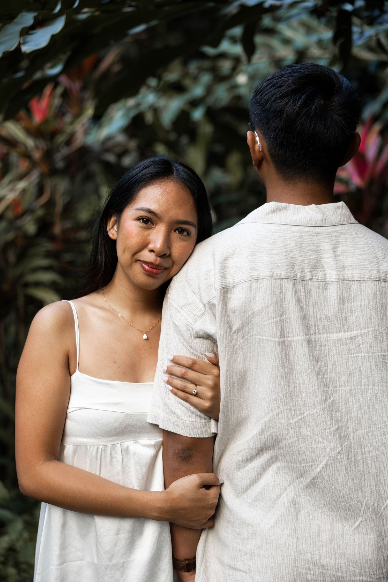 A couple dressed in white poses together against verdant tropical foliage.
