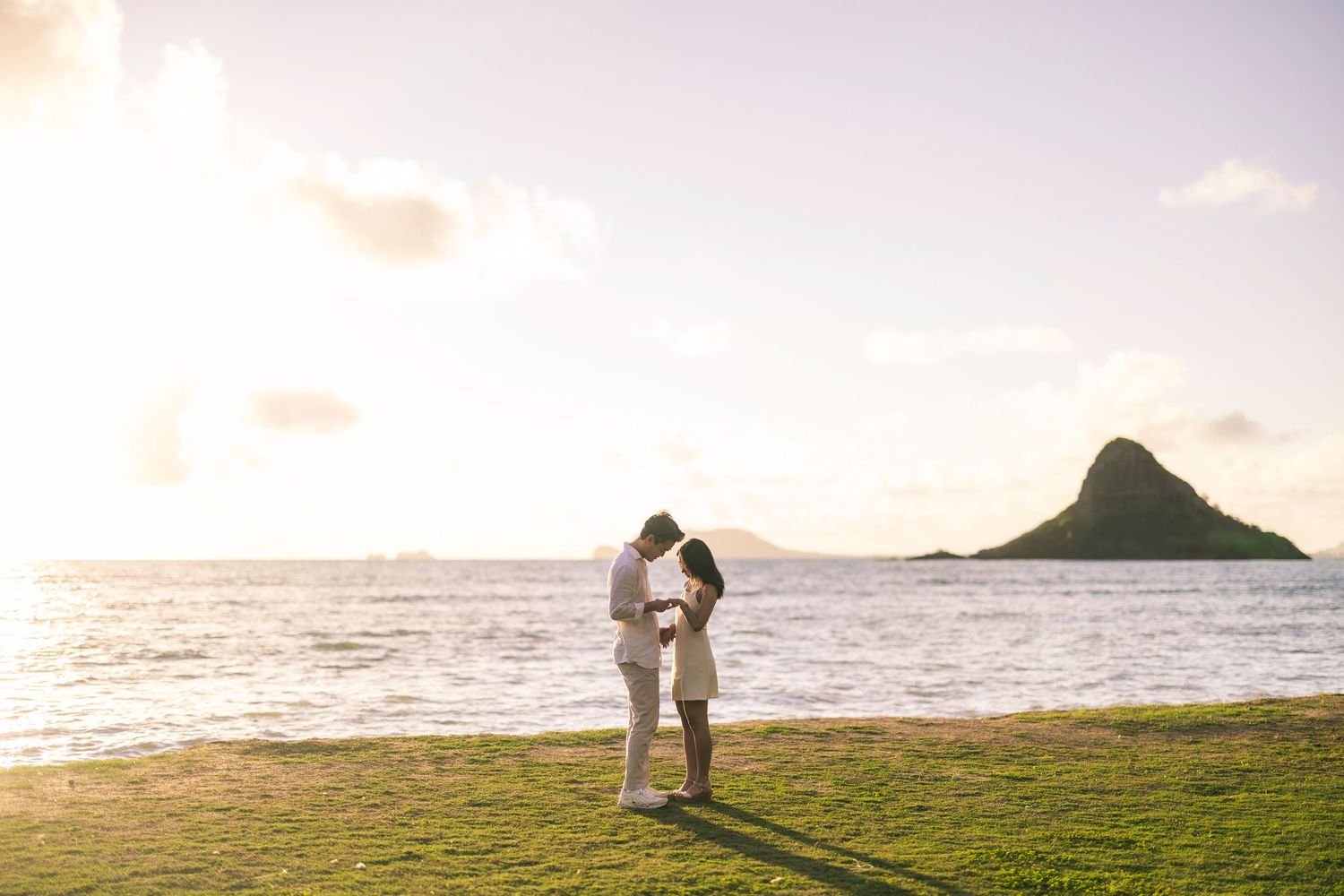 Two people embrace on grassy shore at sunset with Chinaman's Hat island visible in background at Kualoa Beach, Hawaii.