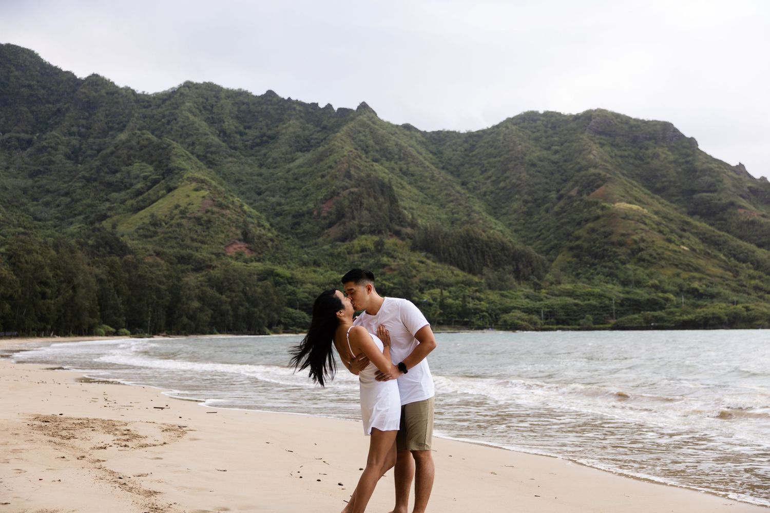 A couple enjoys a romantic walk along a scenic beach with lush green mountains in the background.