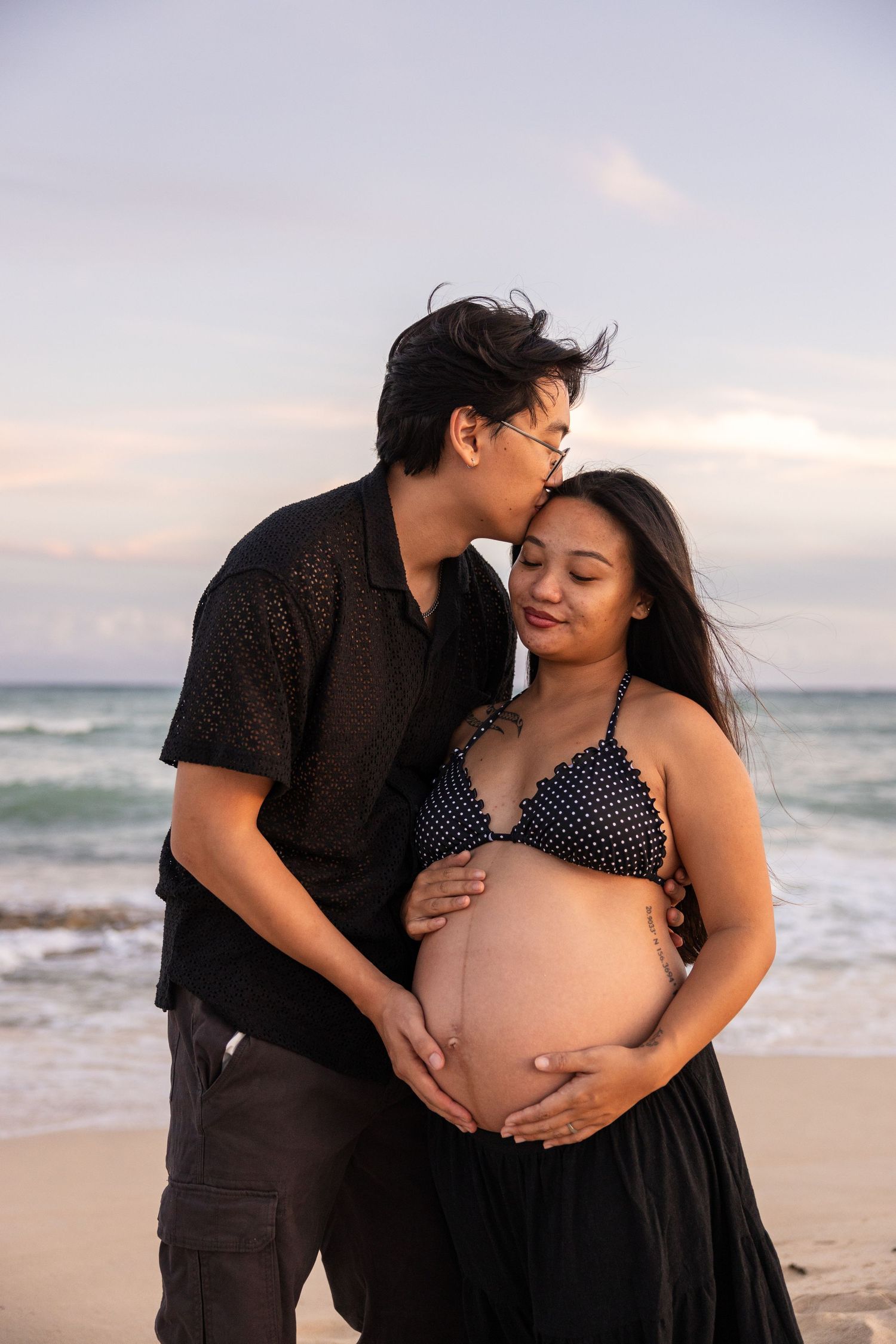 Couple shares intimate moments during maternity photoshoot on beach at sunset with ocean waves in background.