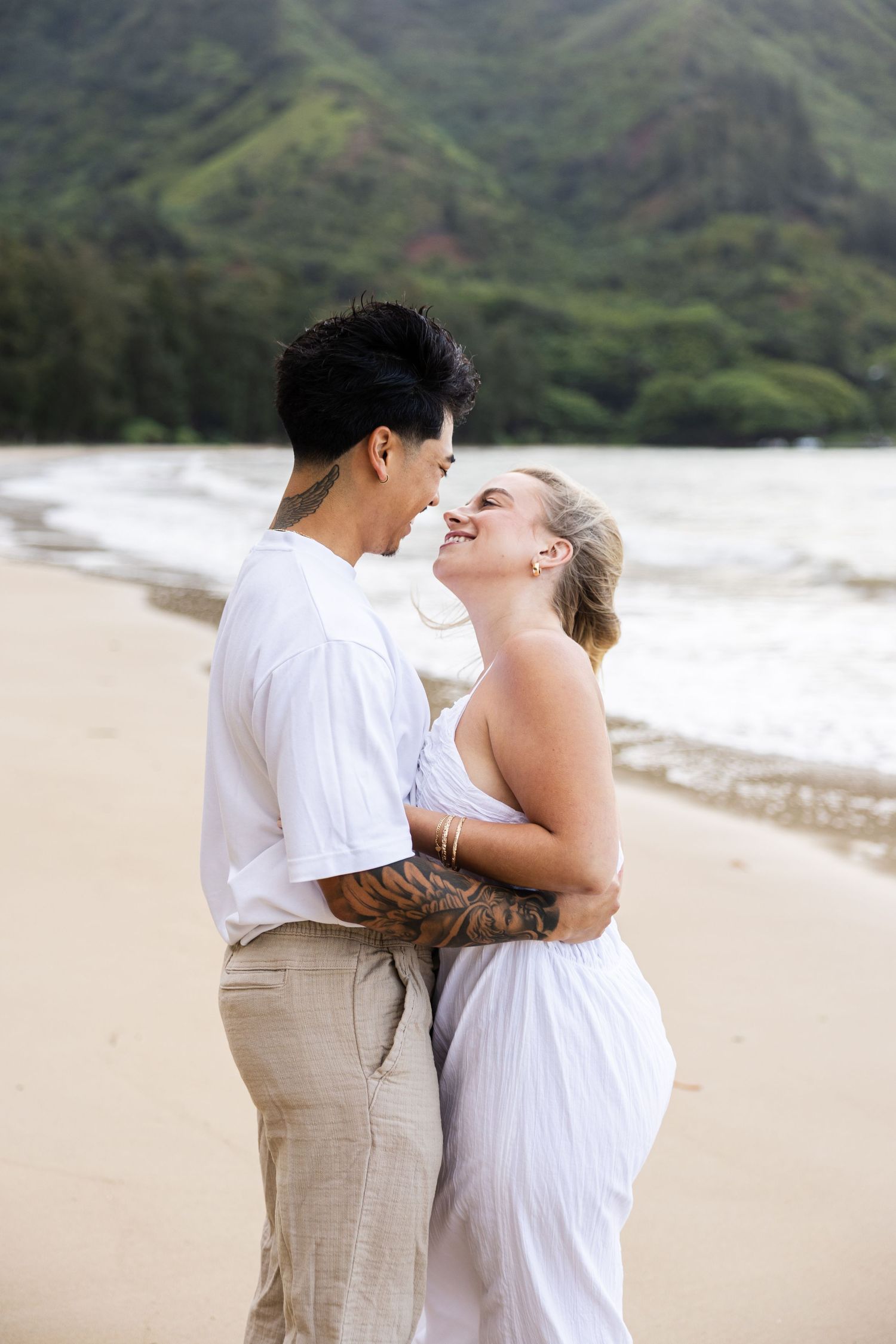 A couple shares a romantic moment together on a scenic beach with lush green mountains in the background.