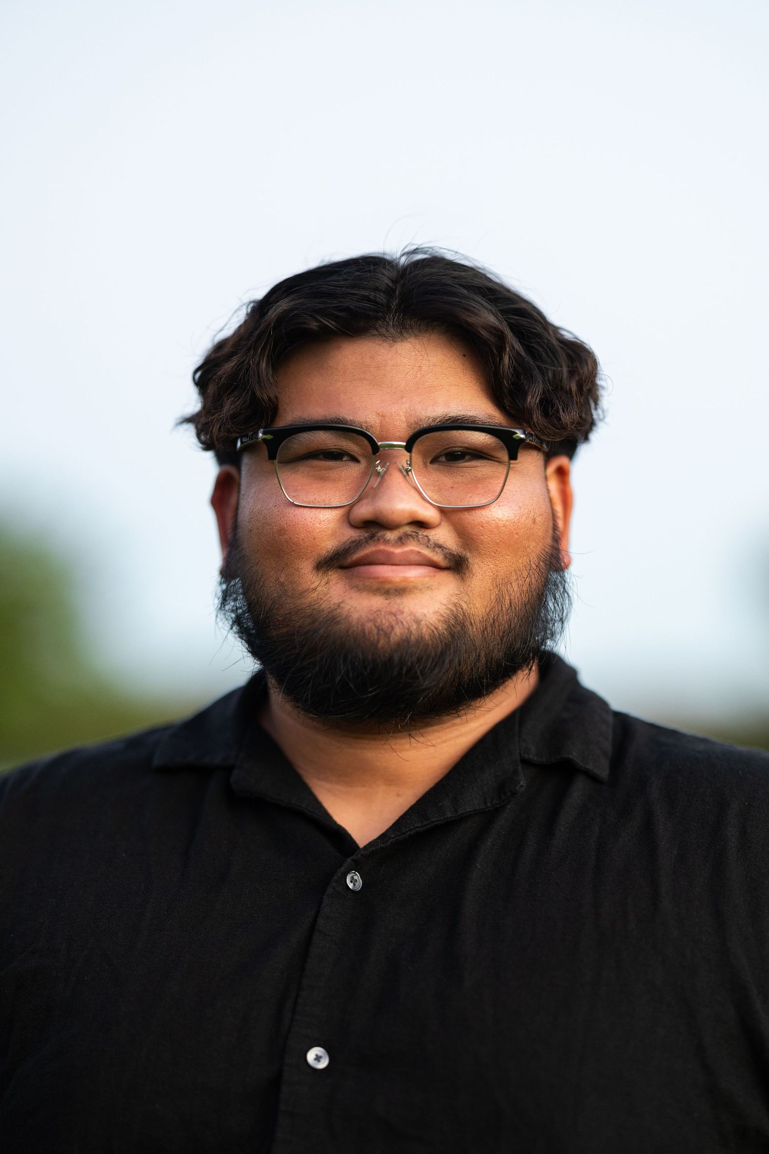 Portrait of a person wearing glasses and a black shirt against a blurred outdoor background.