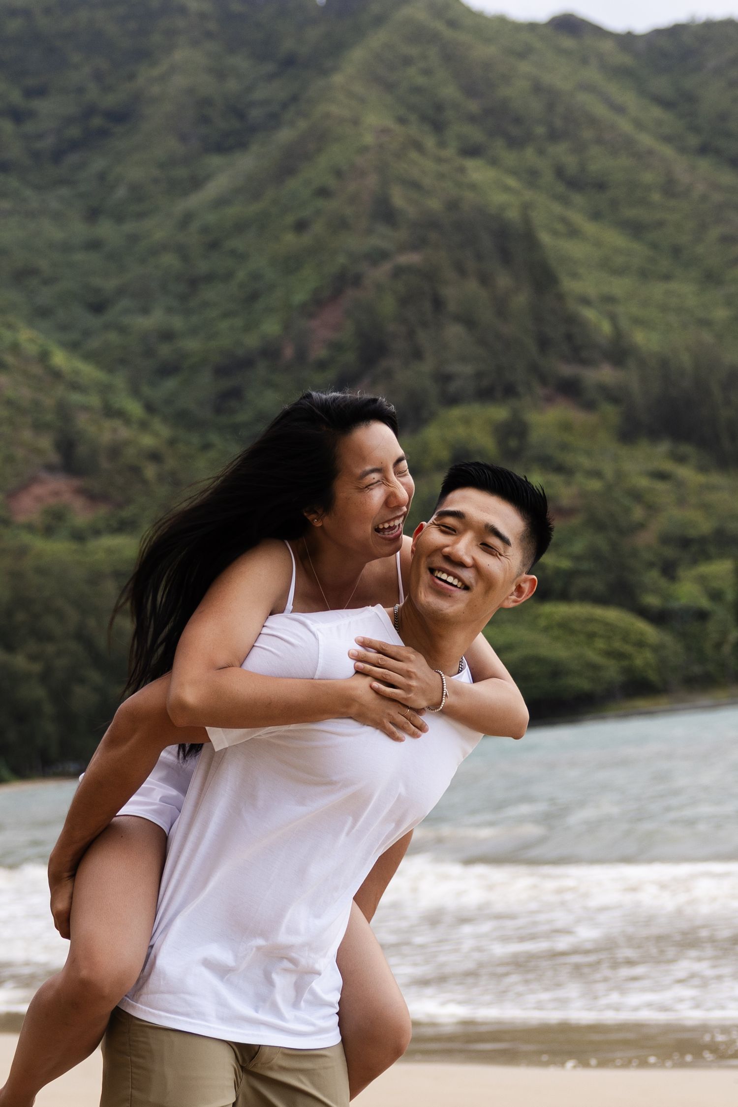 A romantic couple shares joyful laughter and embraces on a scenic beach with lush mountains in the background.
